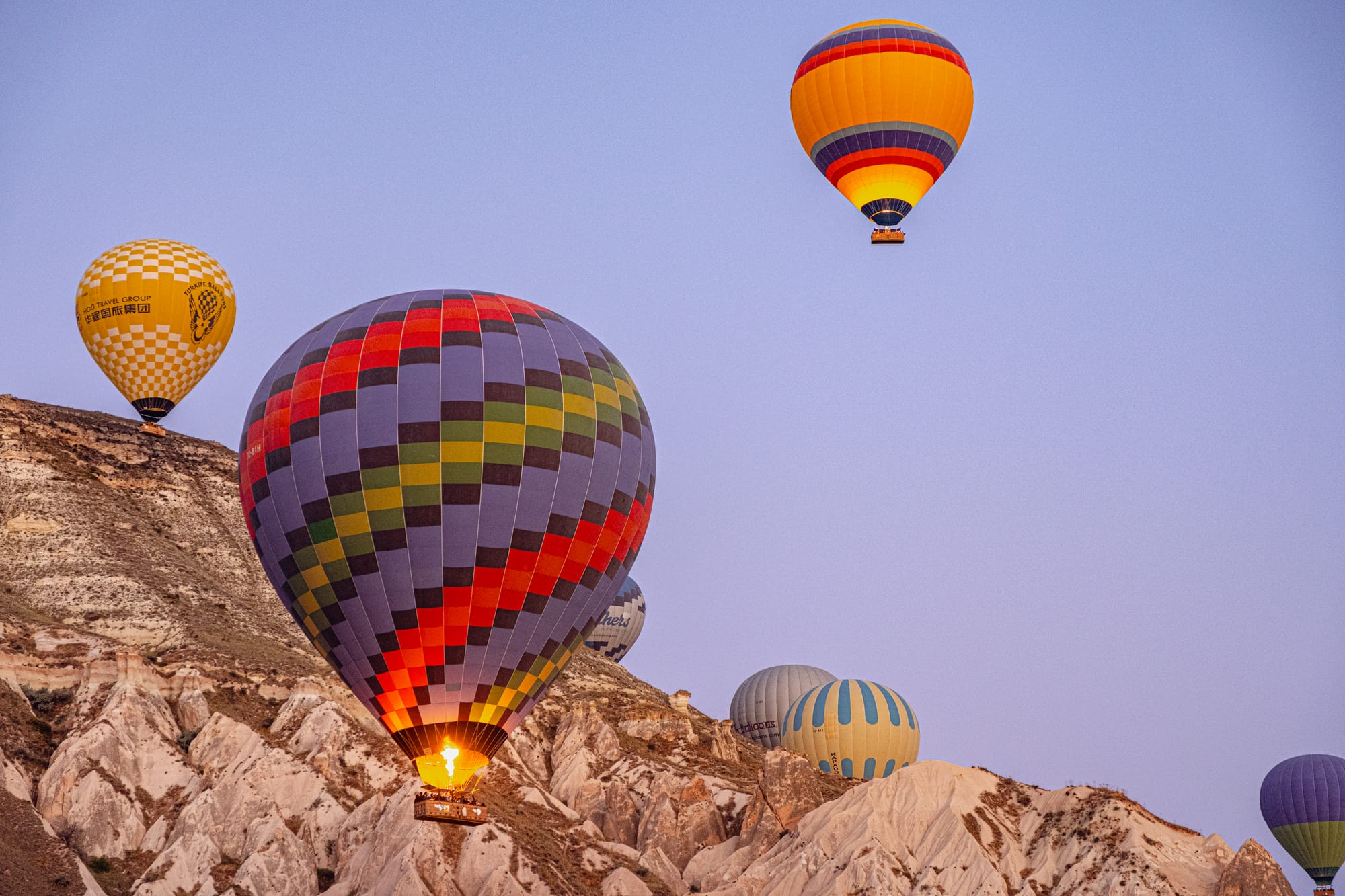 Hot air balloons in Cappadocia at sunrise, including a large multi-colored checkered balloon with its burner lit, floating above rugged rock formations, with other balloons in yellow, blue, and striped patterns in the background against a clear sky