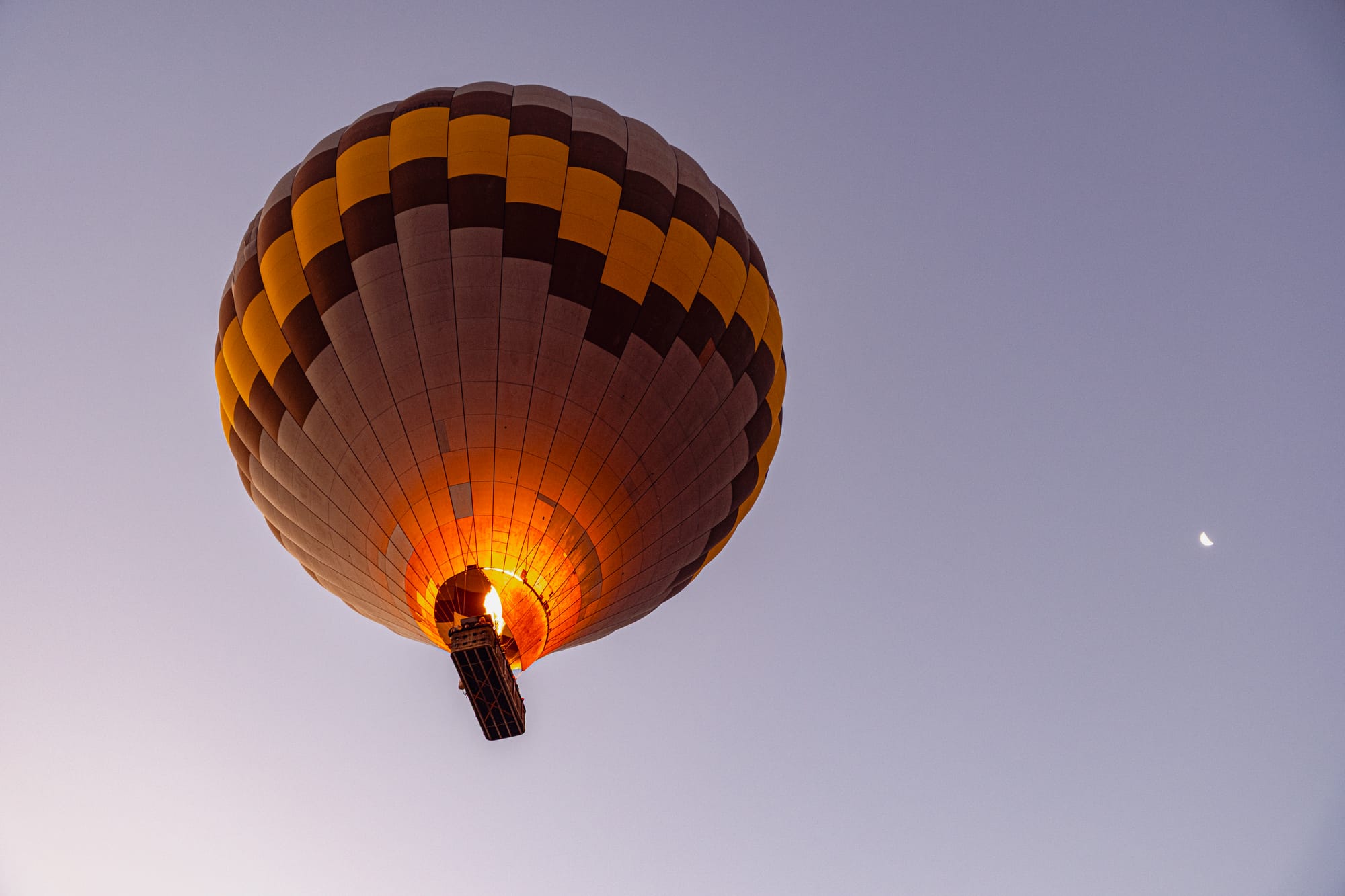 Hot air balloon with yellow and black checkered pattern flying at sunrise in Cappadocia, with the burner flame visible and a crescent moon in the background