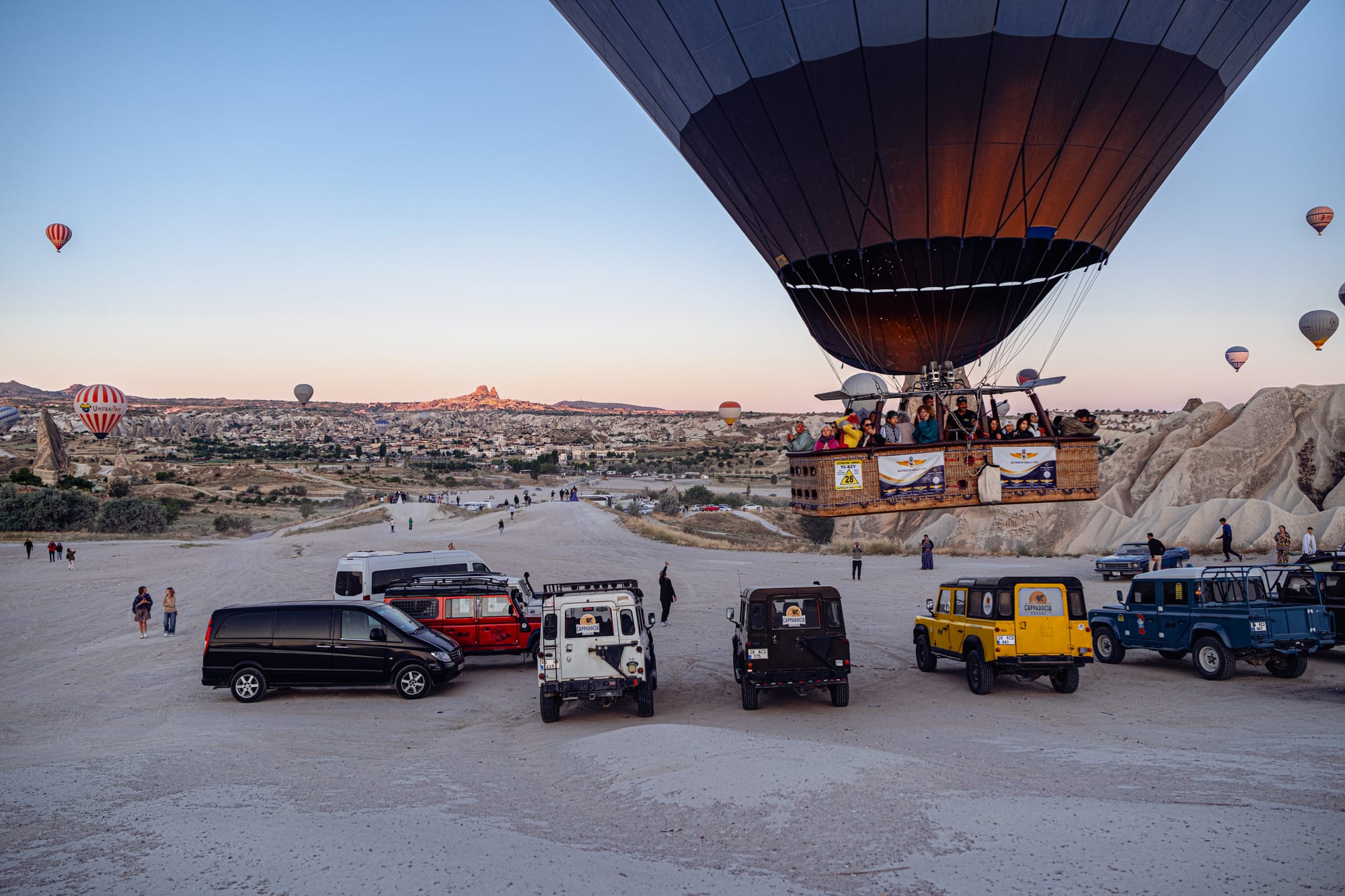 Hot air balloon filled with passengers lifting off near a lineup of jeeps and vans in Cappadocia at sunrise, with the valley, distant rock formations, and other balloons visible in the background