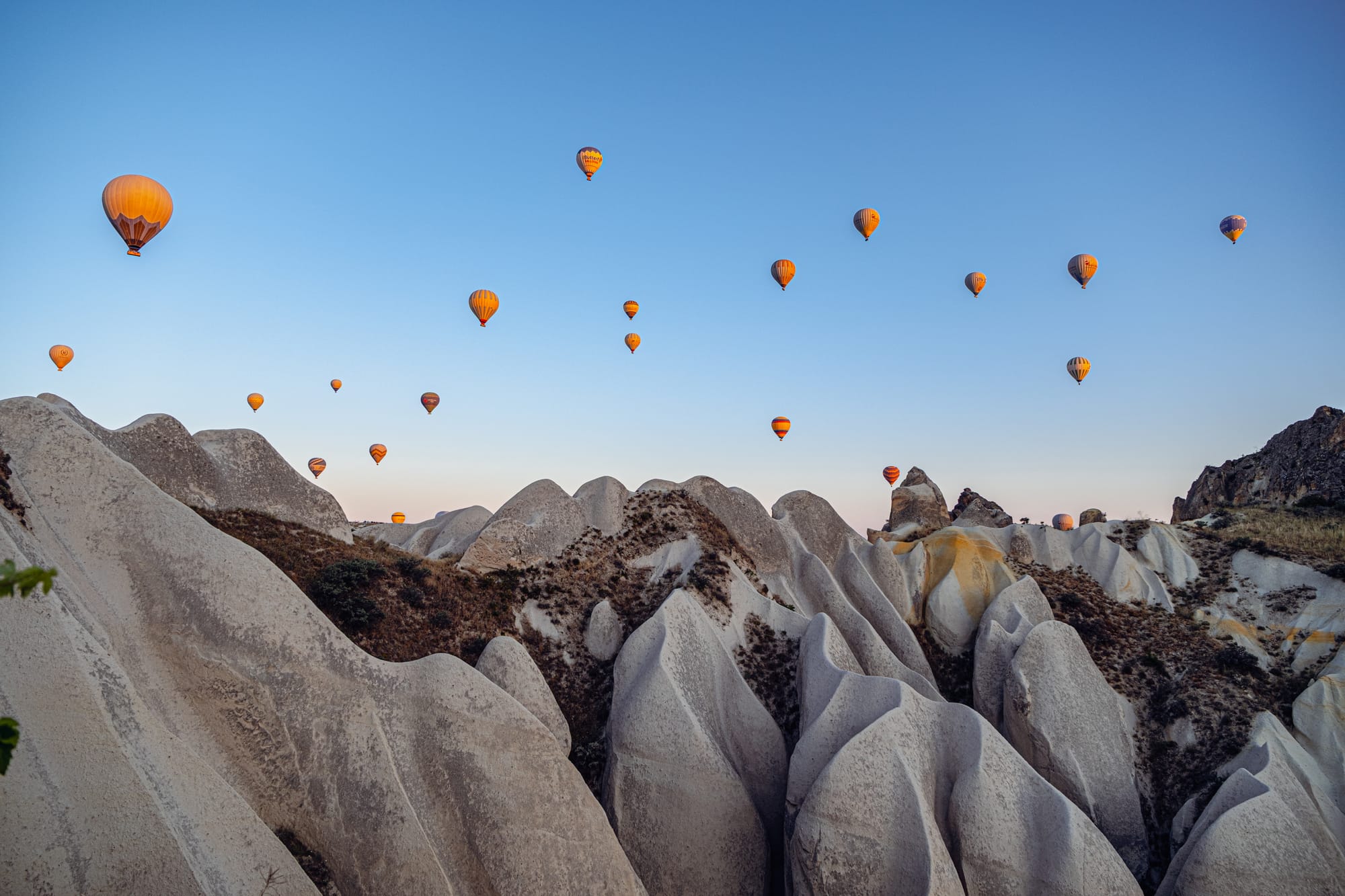Dozens of hot air balloons floating over Cappadocia’s unique white rock formations at sunrise, set against a clear blue sky