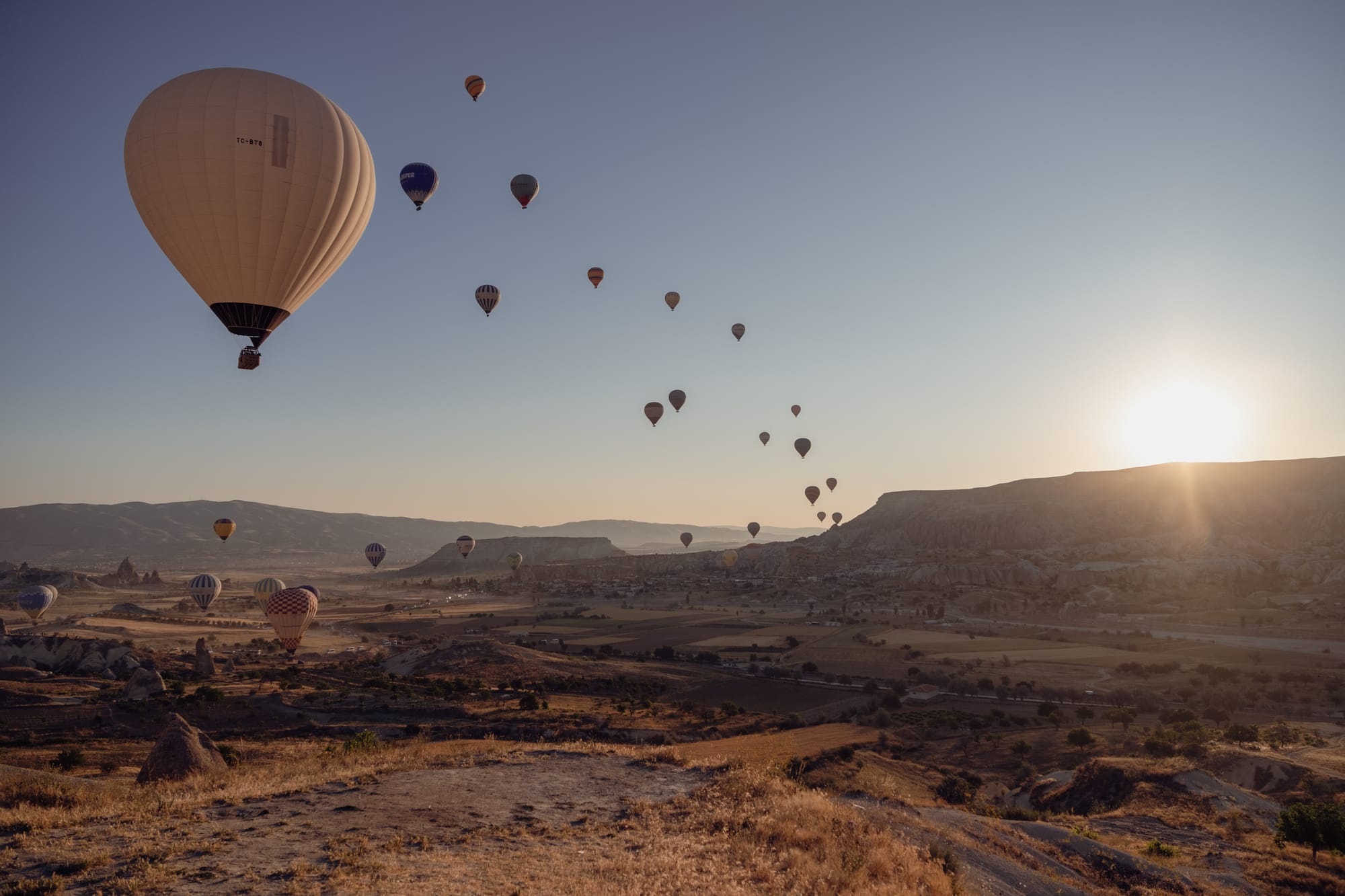 Wide view of Cappadocia at sunrise with numerous hot air balloons floating in the sky, the largest balloon in the foreground, and golden light illuminating the valleys and distant hills
