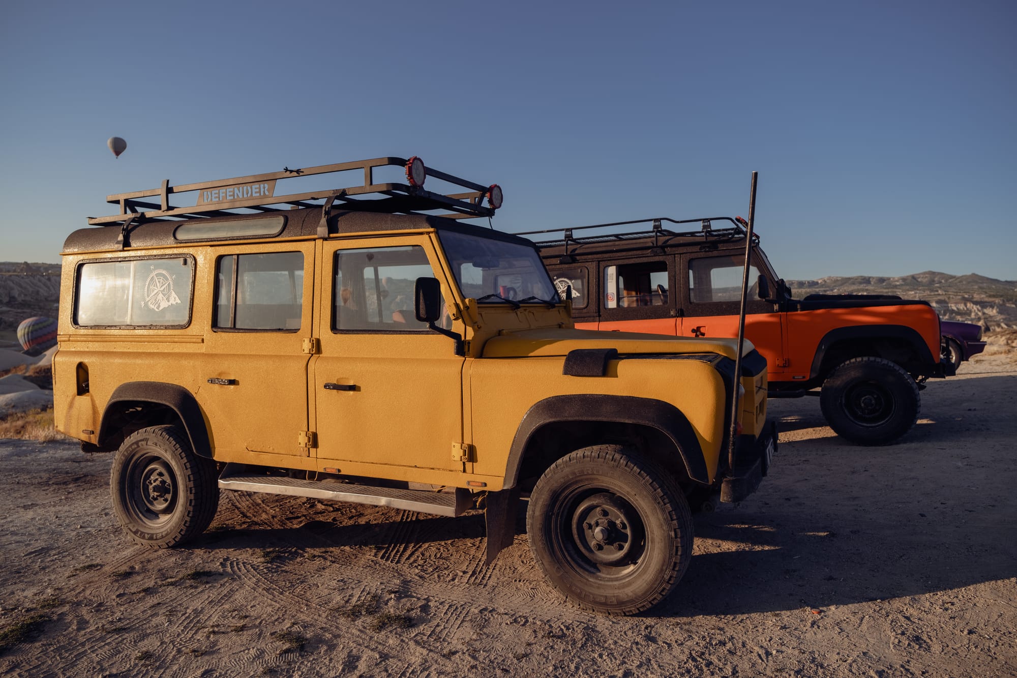 Yellow and orange Land Rover Defender jeeps parked on a dirt overlook in Cappadocia at sunrise, with hot air balloons in the background and mountains on the horizon