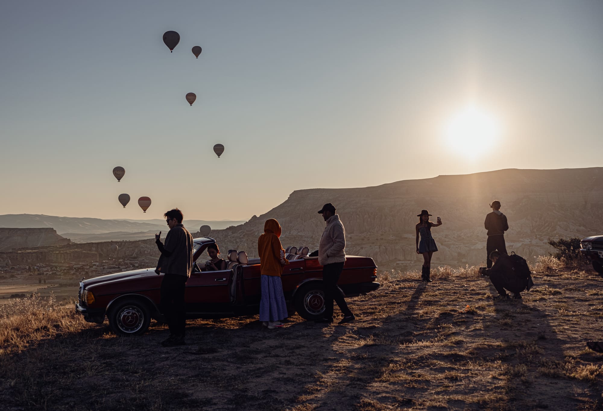 Group of people with a red convertible car on a hilltop in Cappadocia at sunrise, photographing and watching hot air balloons in the sky