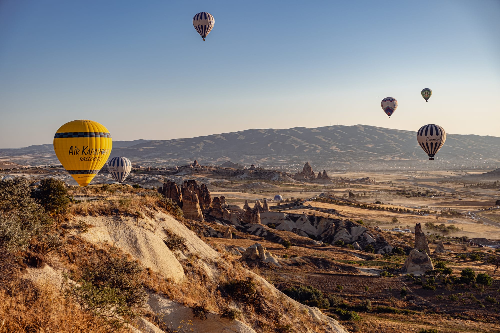 Multiple hot air balloons floating over Cappadocia’s rocky landscape and valleys in the early morning light, with a prominent yellow and blue balloon in the foreground and distant mountains in the background