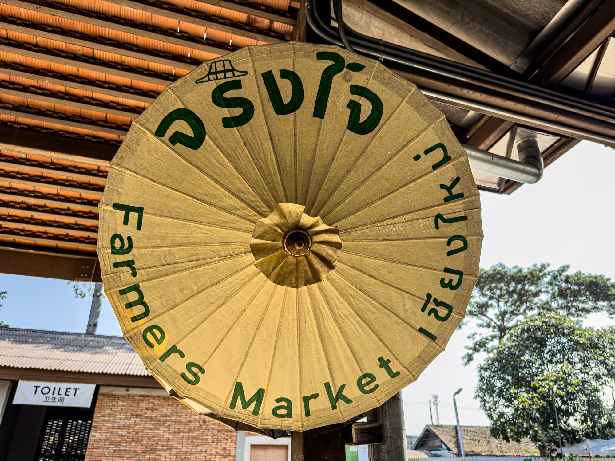 Close-up of a large yellow umbrella with green lettering reading “Farmers Market Chiang Mai” in English and Thai, symbolizing the Jing Jai Market entrance