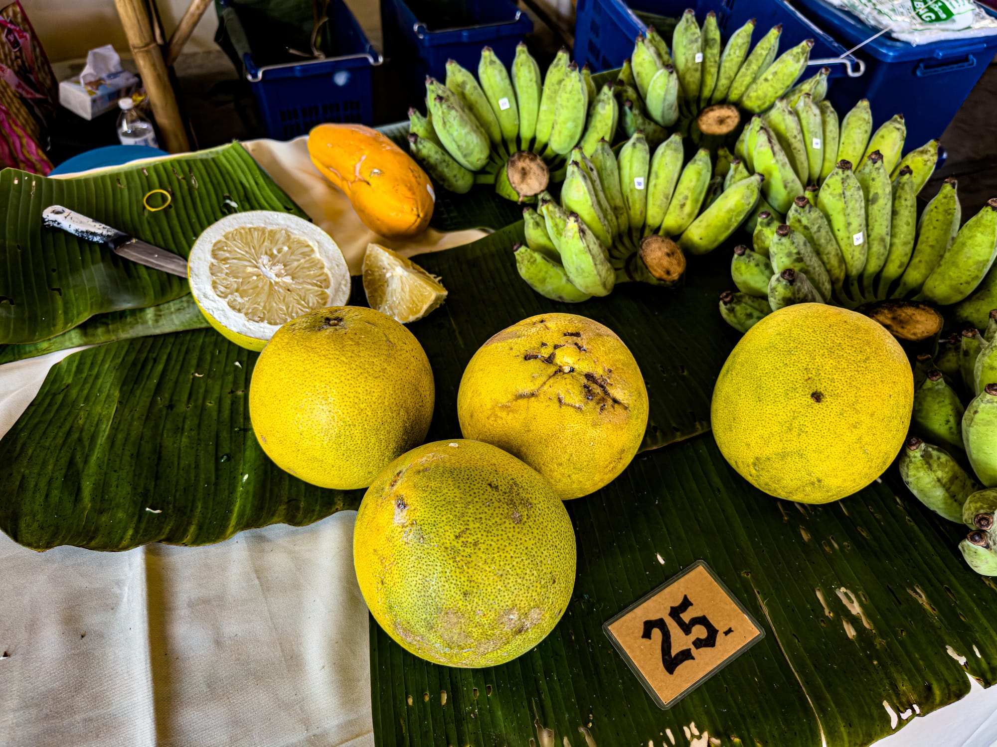 Close-up of a fruit stall at Jing Jai Market showing whole and halved pomelos, bunches of green bananas, and a ripe papaya arranged on banana leaves with a price tag of 25 baht