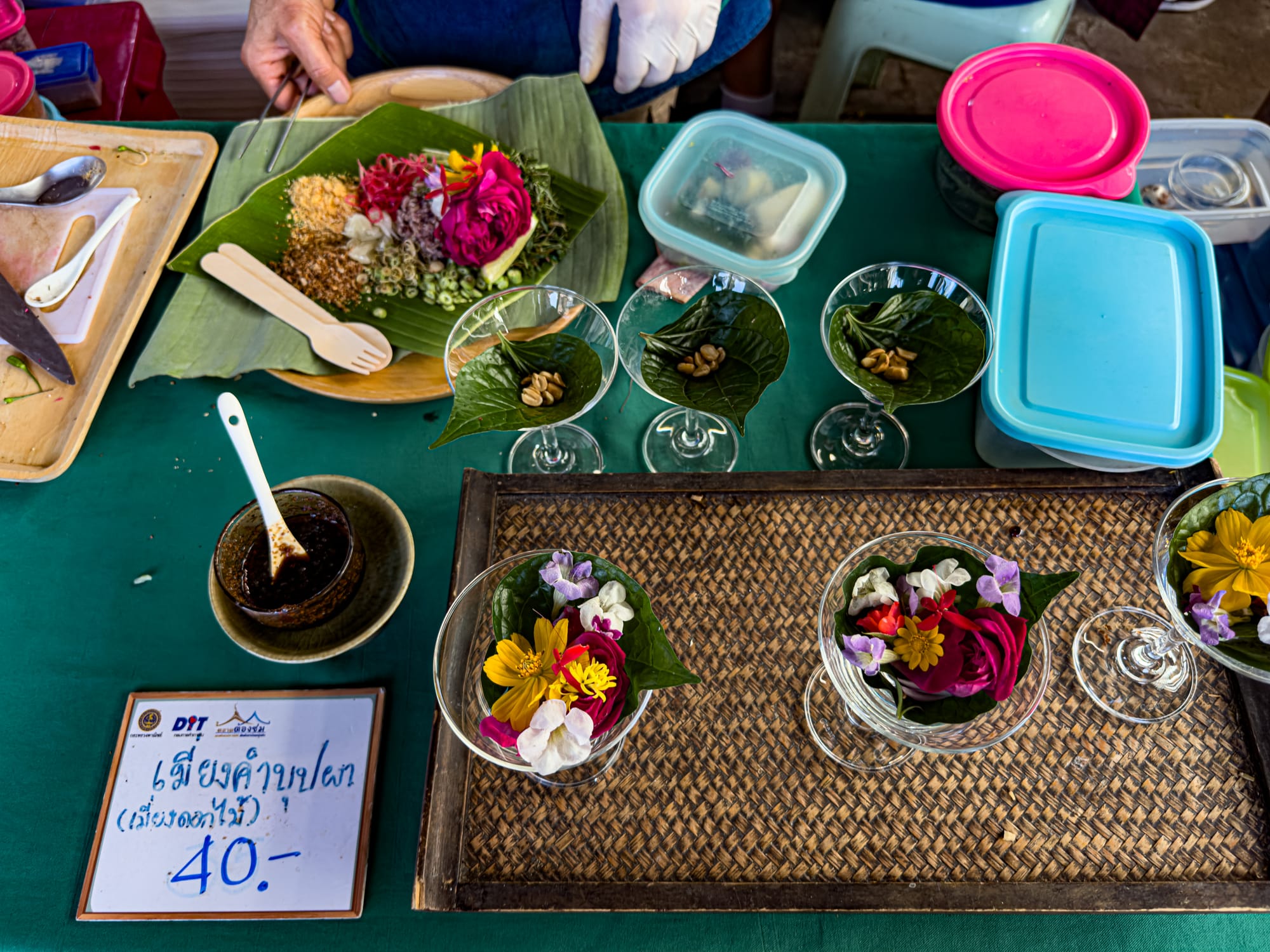 Close-up of a vendor’s stall at Jing Jai Market displaying colorful edible flower salads in glass cups, with petals, herbs, and peanuts arranged on betel leaves, alongside a banana leaf platter of ingredients and a sign showing the price of 40 baht