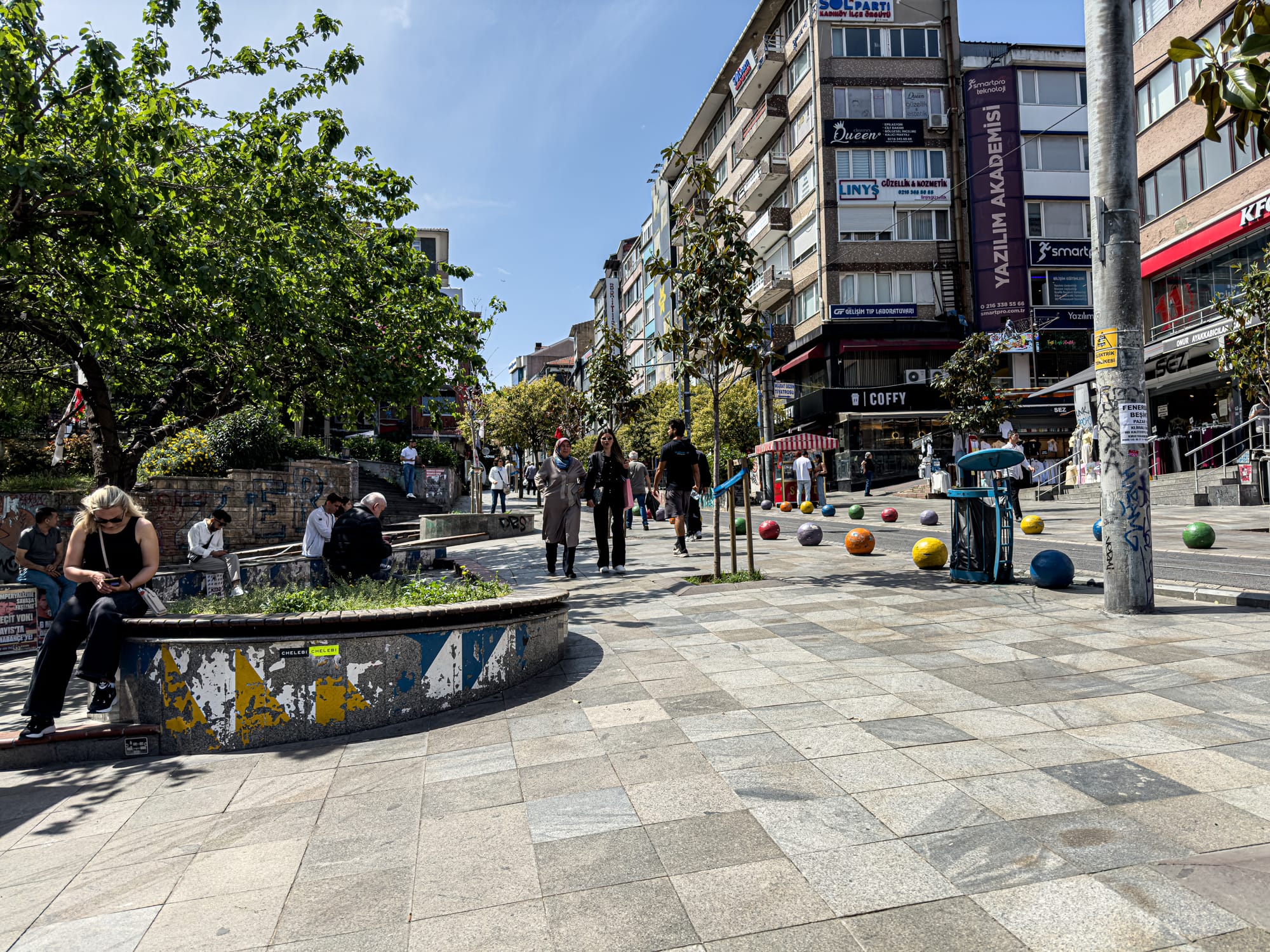 Urban square in Kadıköy with colorful bollards, people sitting on benches, and shops lining the street on a sunny day