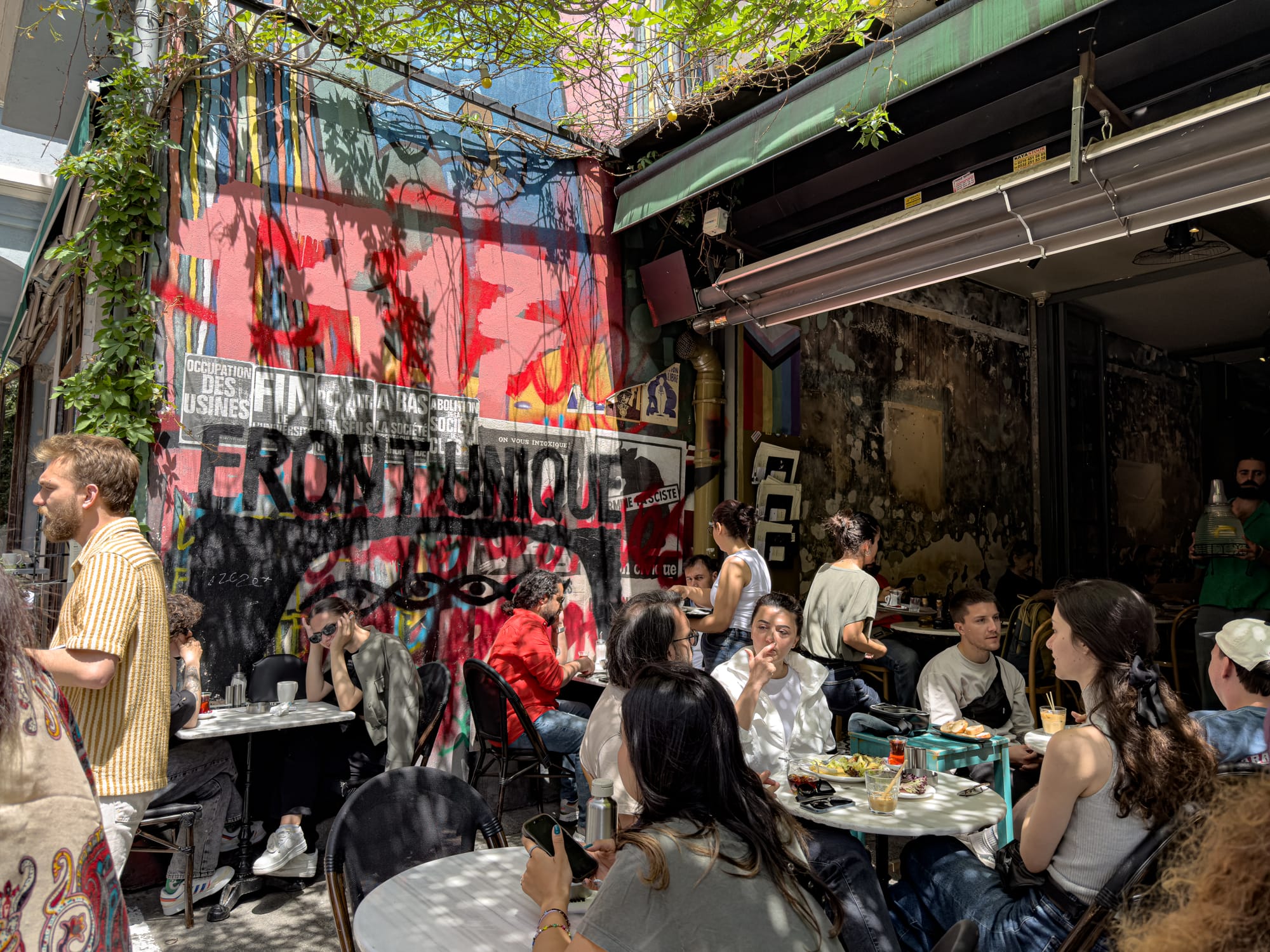 Crowded courtyard café in Kadıköy with groups of people sitting at small tables, surrounded by colorful graffiti, wall art, and dappled sunlight filtering through leafy vines