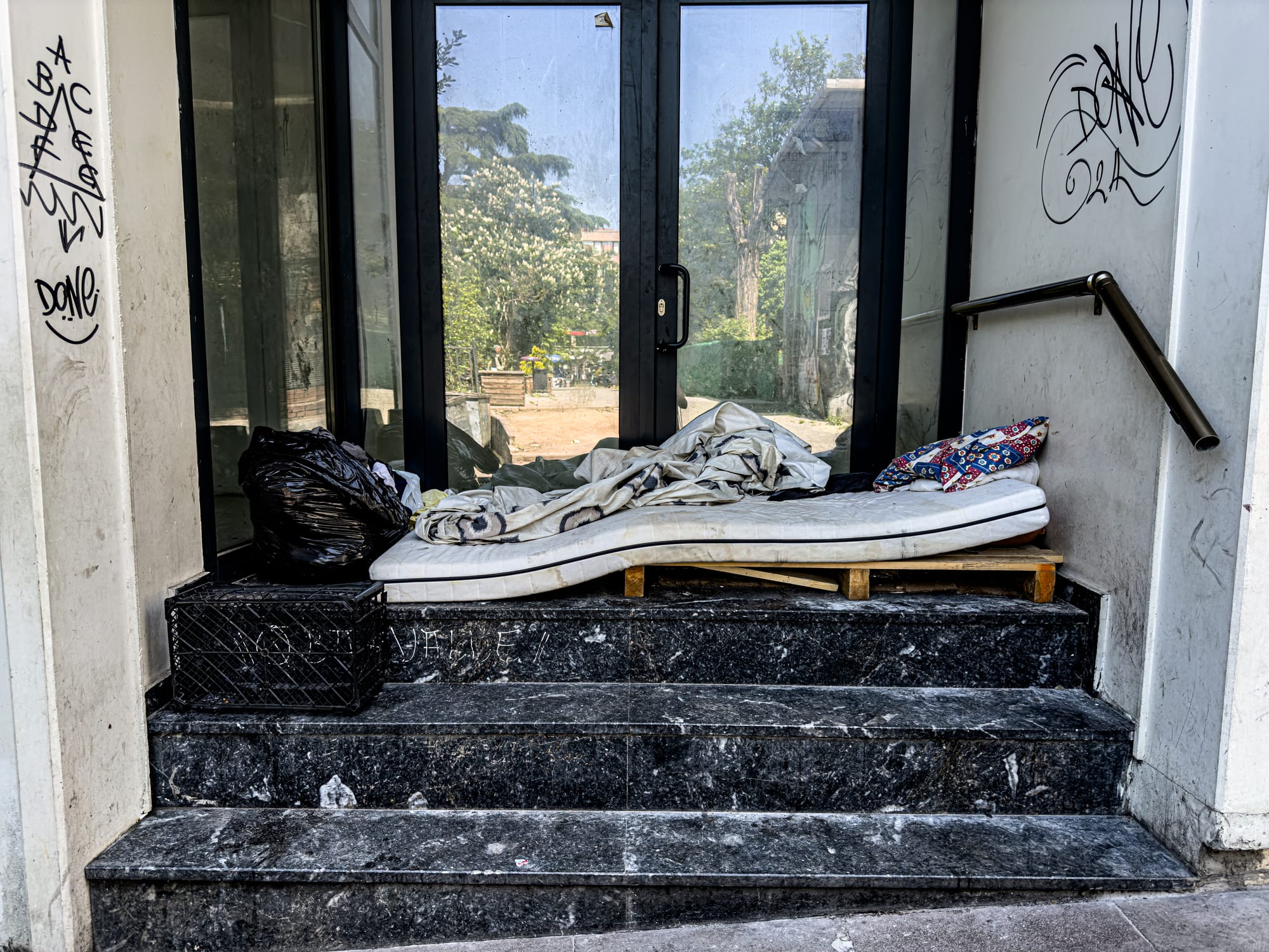 A street-level photo of a thin mattress with sheets and a patterned pillow placed on a wooden pallet in a doorway, flanked by graffiti-covered walls and a black garbage bag—suggesting a temporary shelter for someone unhoused in Kadıköy