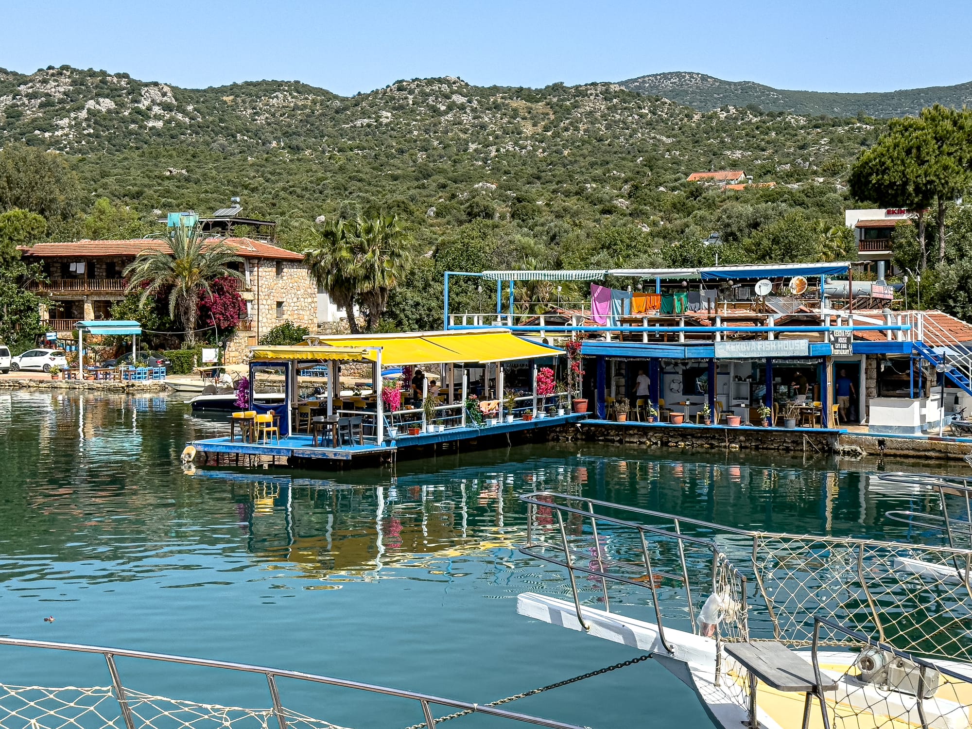 Colorful waterfront with docked boats and floating cafés at the Kekova departure point, framed by green hills and traditional stone houses