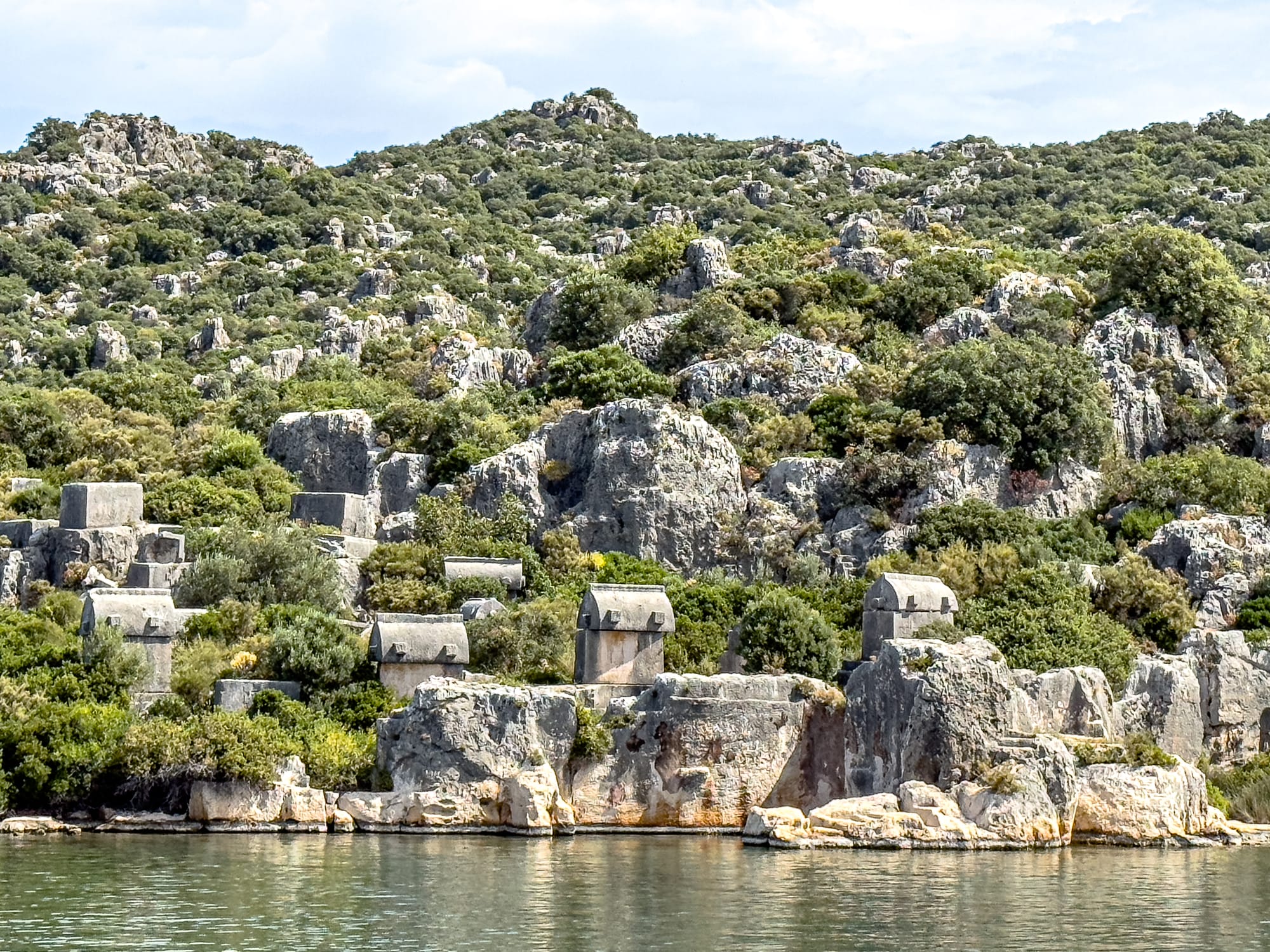 Dozens of ancient Lycian sarcophagi scattered across a rocky hillside near the water’s edge at Kekova, Turkey