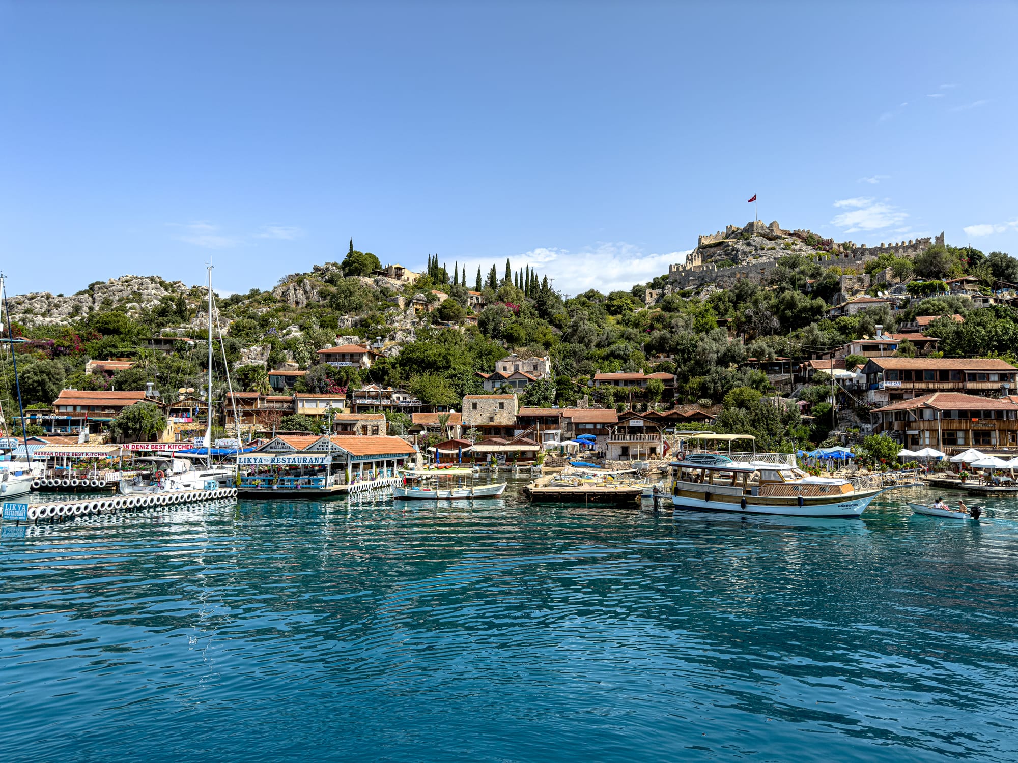Waterfront view of Kaleköy village near Kekova, with docked boats, stone houses, and a hilltop Byzantine castle flying the Turkish flag