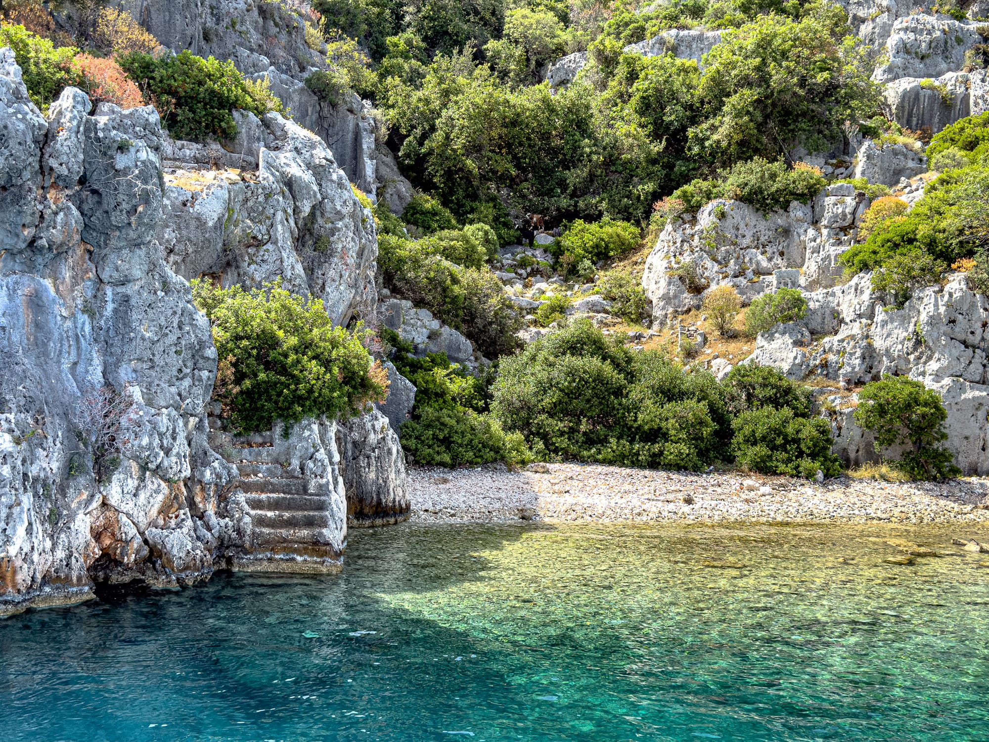 Ancient stone staircase carved into a cliffside at Kekova, Turkey, leading directly into the sea beside a small pebble beach and rocky slope