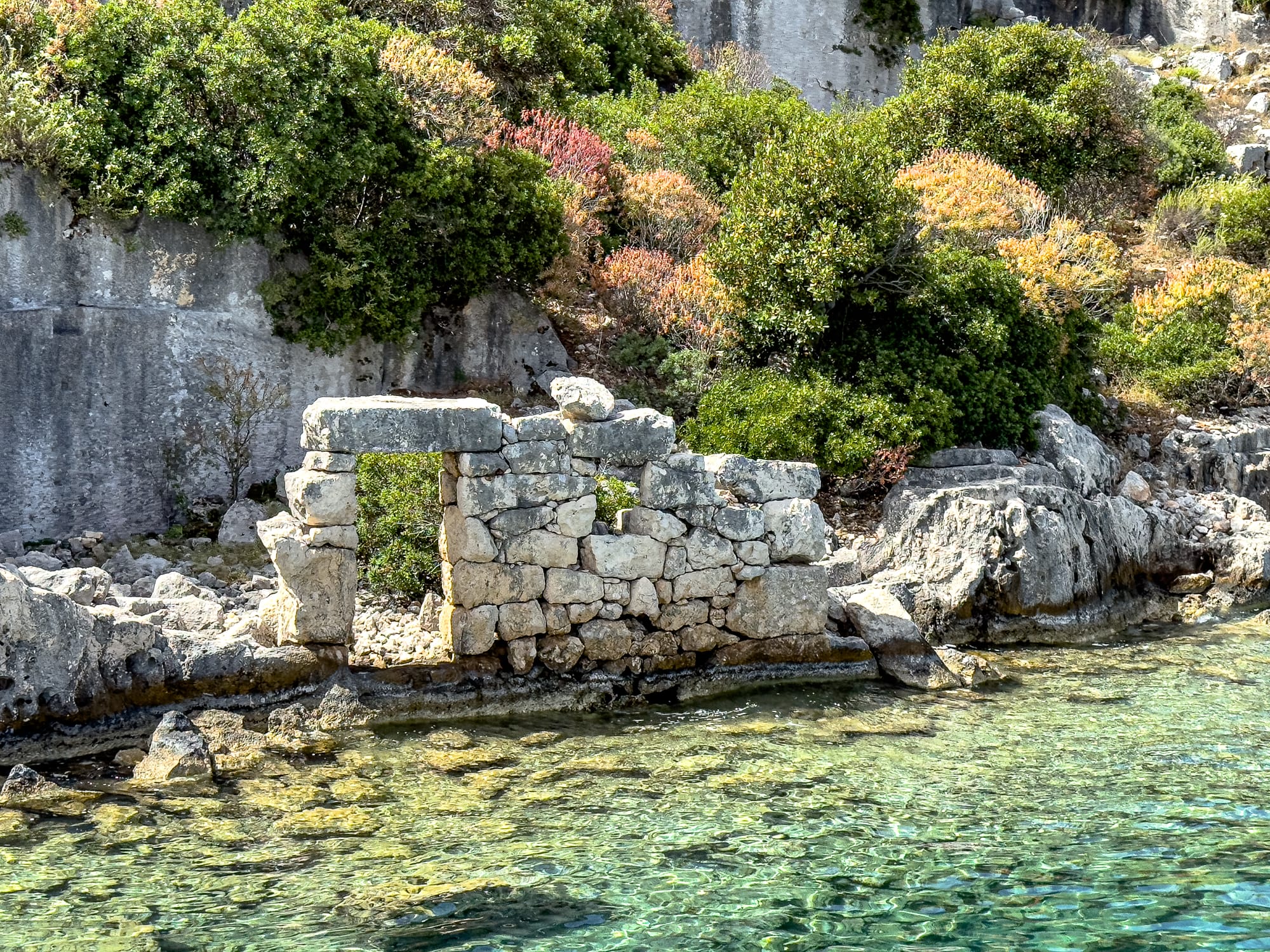 Ruined stone doorway of an ancient building at the water’s edge in the sunken city of Kekova, surrounded by shrubs and rocky shoreline