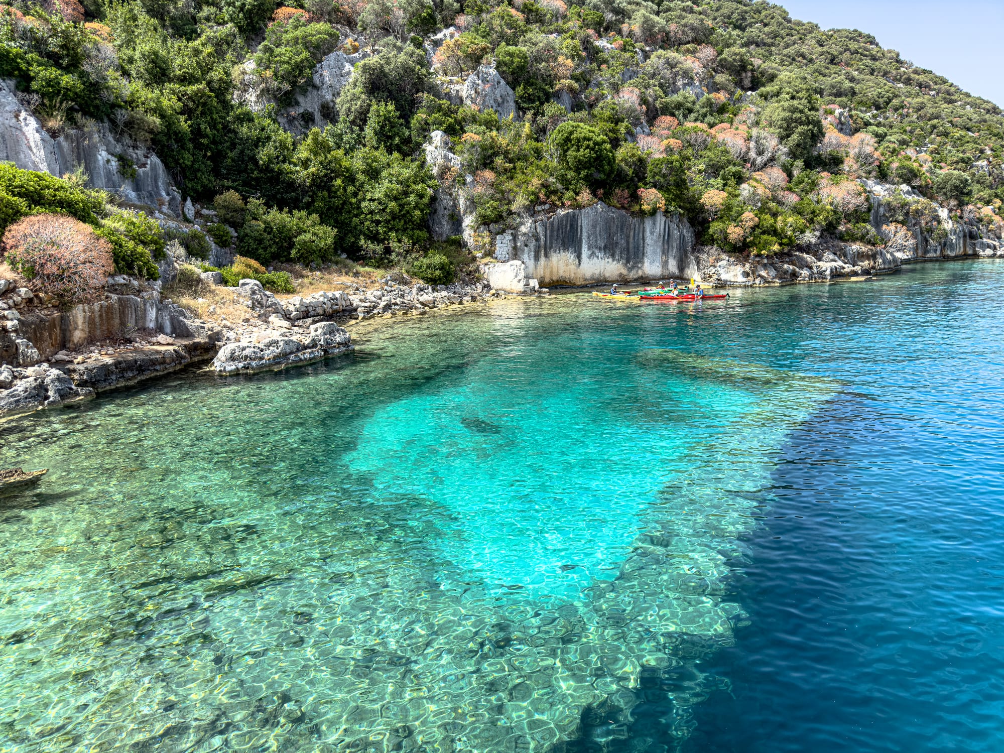 Clear turquoise waters reveal the sunken remains of Kekova’s ancient port, with kayakers paddling past the shoreline ruins above