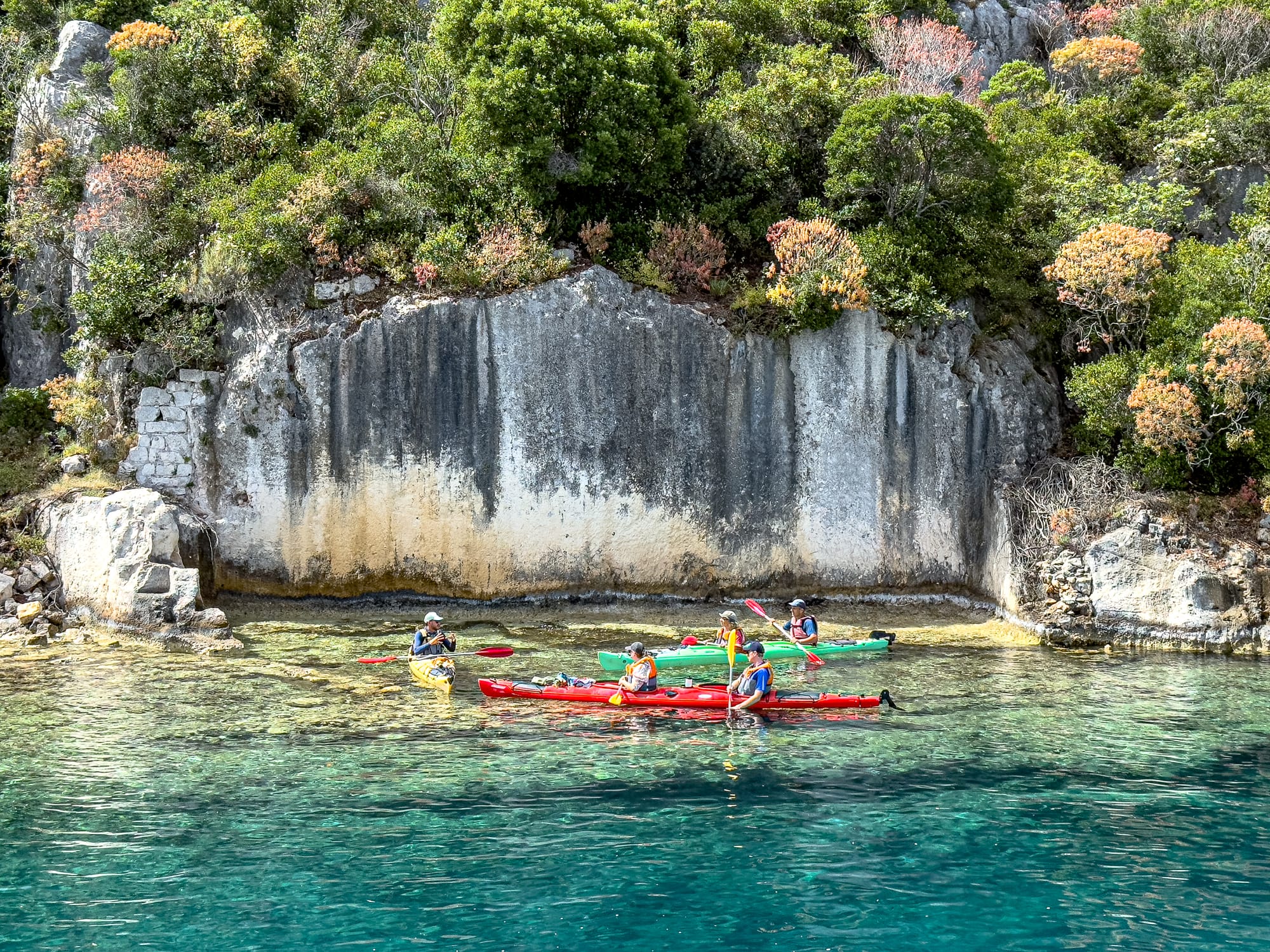 Group of kayakers paddling along the shoreline of Kekova, passing a large stone wall remnant of the sunken Lycian city, partially submerged in clear turquoise water
