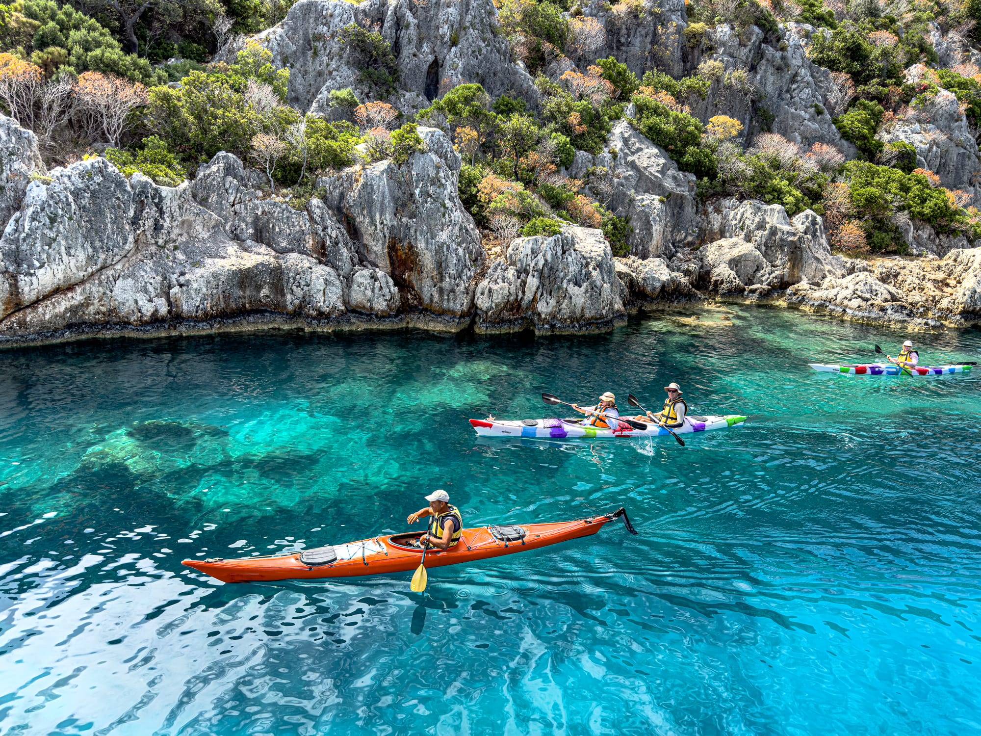 Four kayakers paddle over turquoise waters along the rocky shoreline of Kekova, near the submerged ruins of the ancient Lycian city