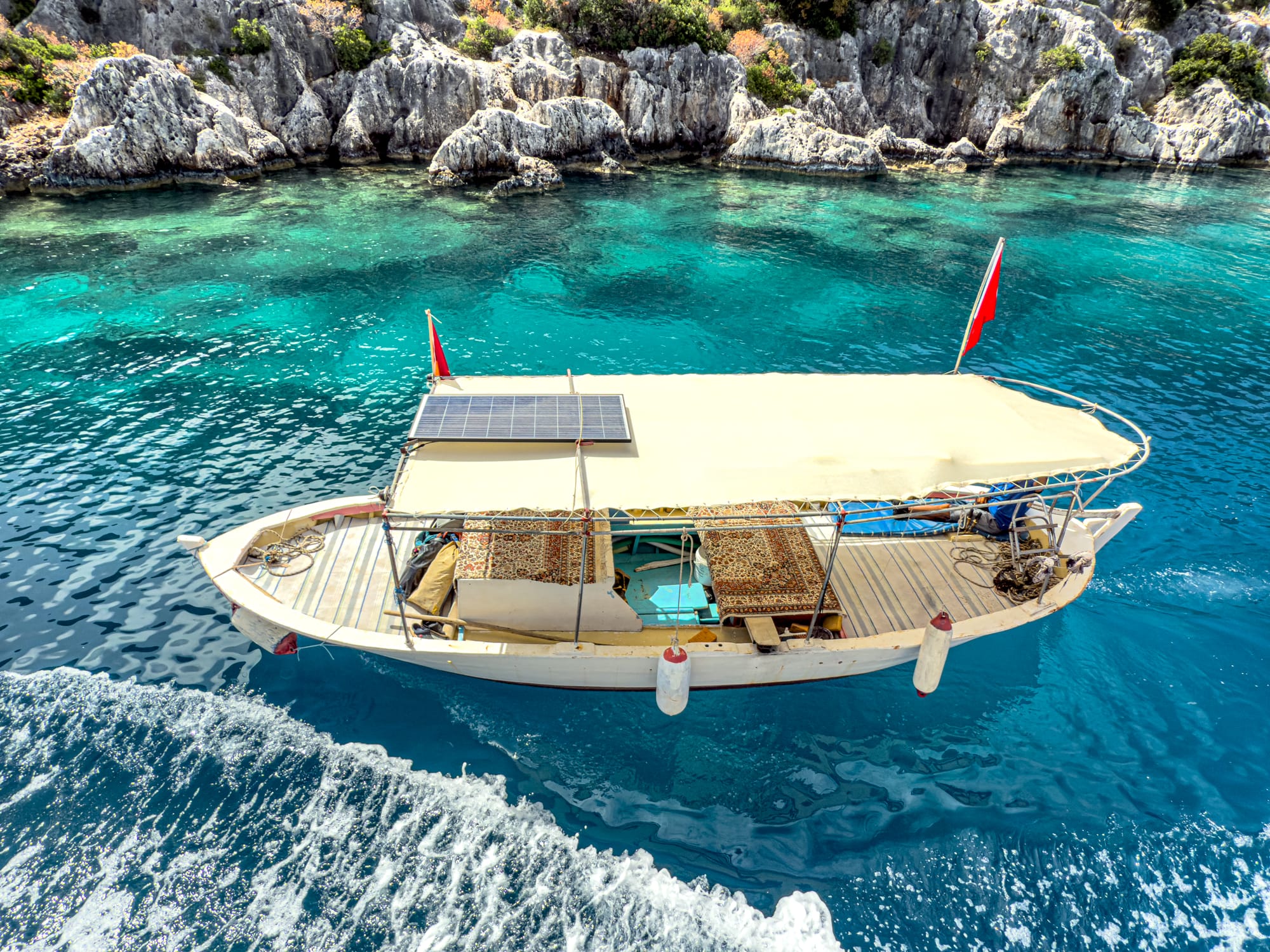 Small Turkish boat with a canopy and carpeted seating cruises through the bright turquoise waters off the coast of Kekova, with rocky shoreline in the background