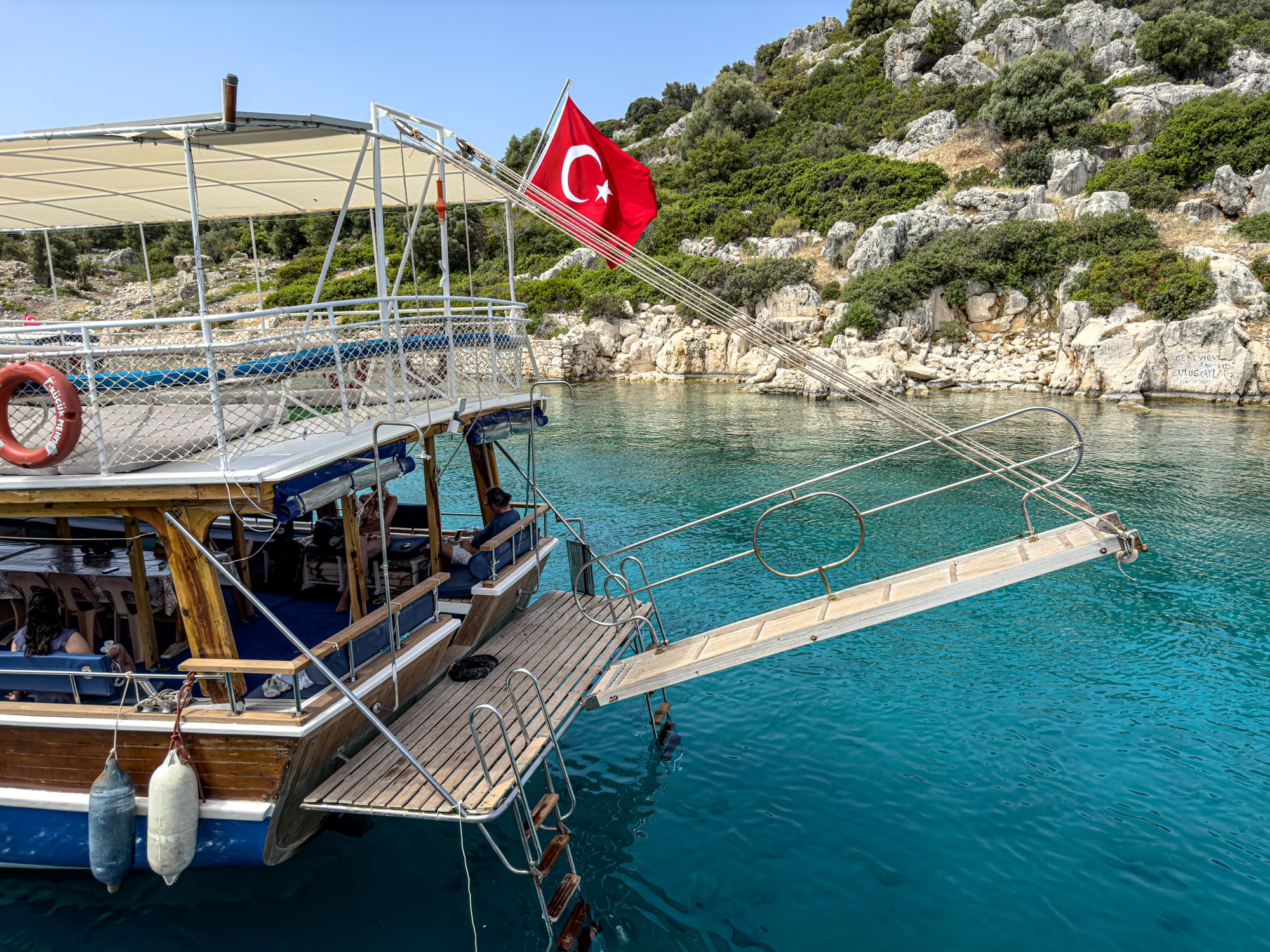 Turkish flag flying from a wooden tour boat anchored in a turquoise bay near Kekova, with a gangway extended over the water and rocky hills in the background