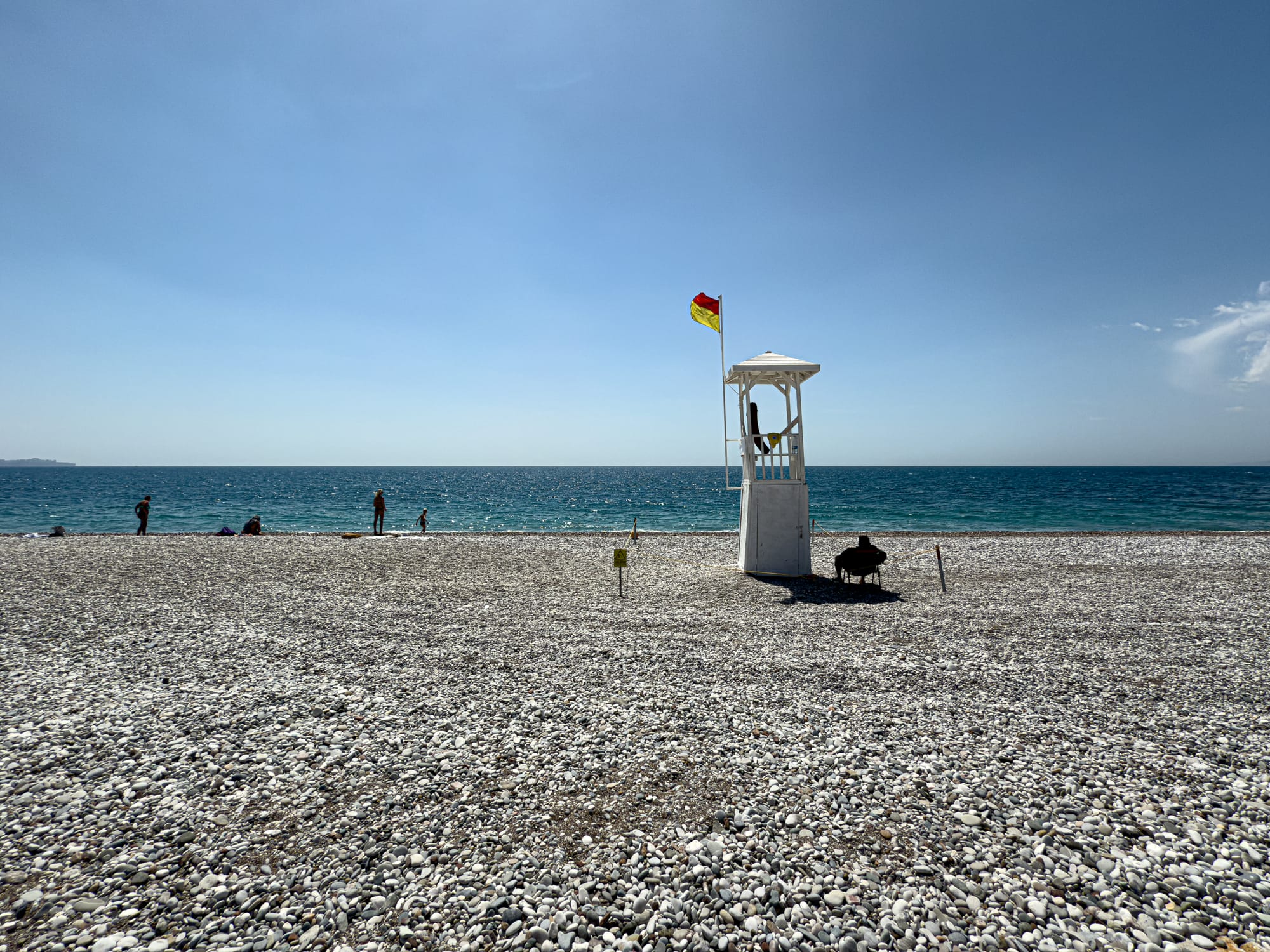A lifeguard tower stands on the rocky stretch of Konyaaltı Beach, facing the turquoise Mediterranean under a wide, cloudless sky