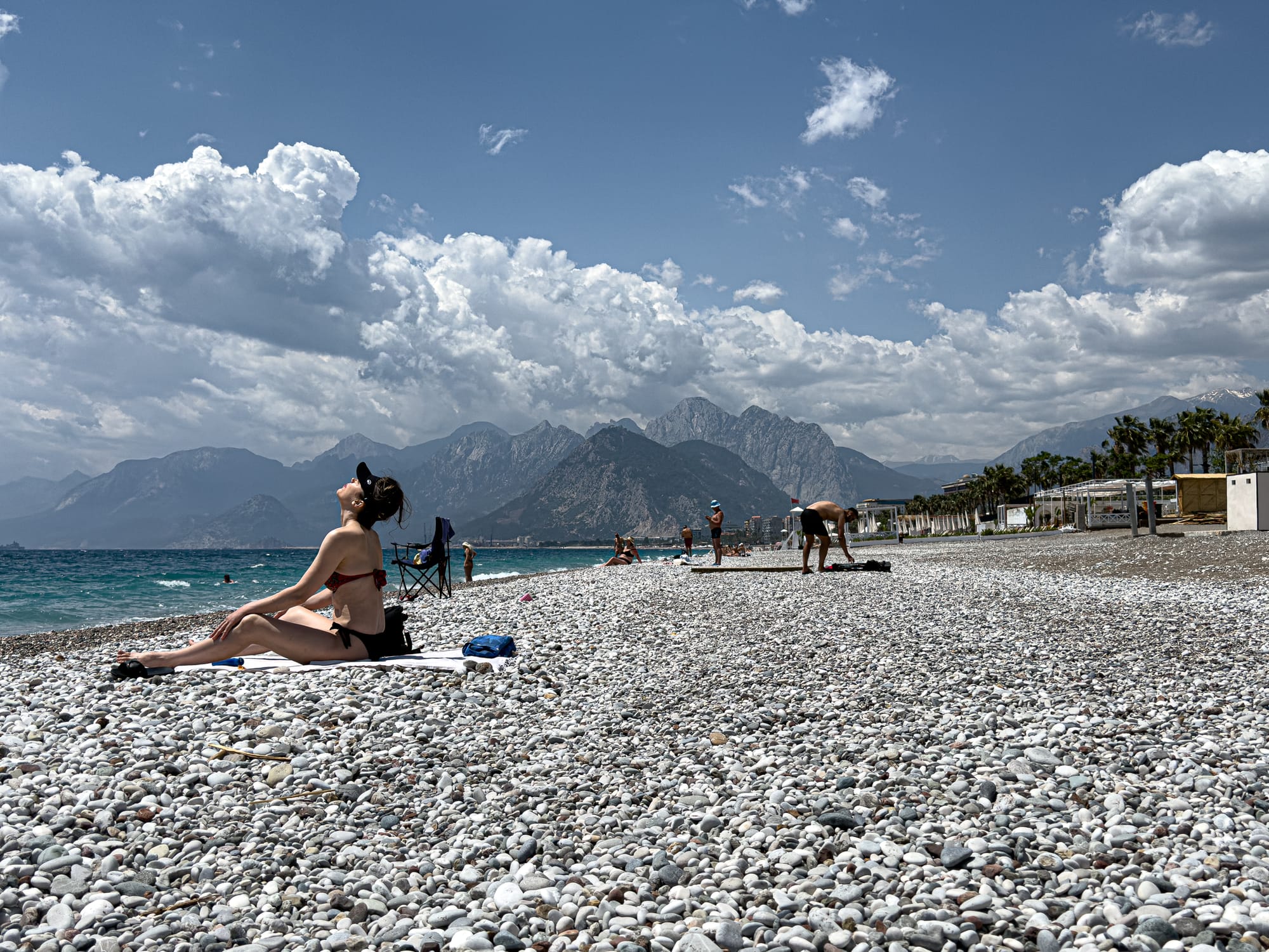 A woman sunbathes on a towel at rocky Konyaaltı Beach, with towering mountains and dramatic clouds in the background