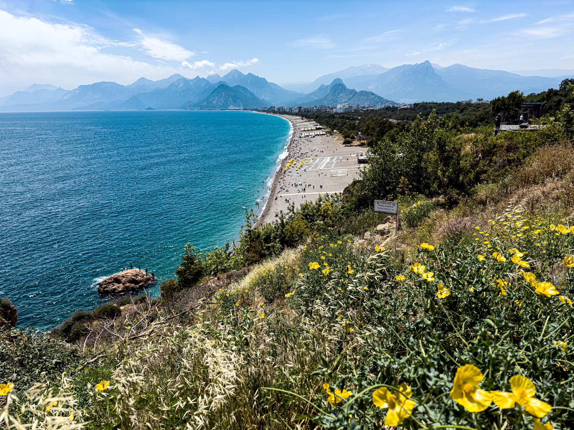 View from a cliffside trail overlooking the long curve of Konyaaltı Beach, with yellow wildflowers in the foreground and dramatic mountains rising beyond the sea