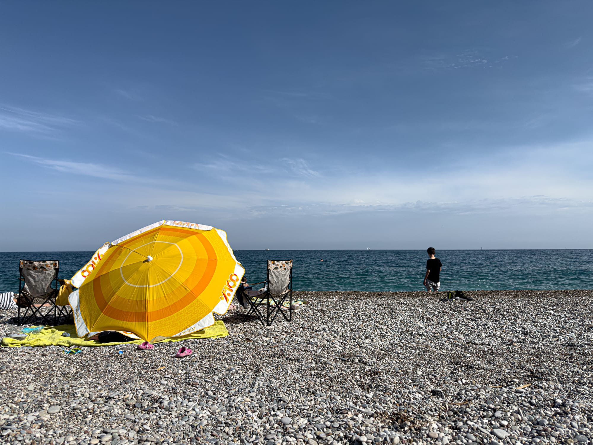 A bright yellow beach umbrella and foldable chairs set up on the rocky shoreline of Konyaaltı Beach, with a person standing at the water’s edge under a wide, open sky