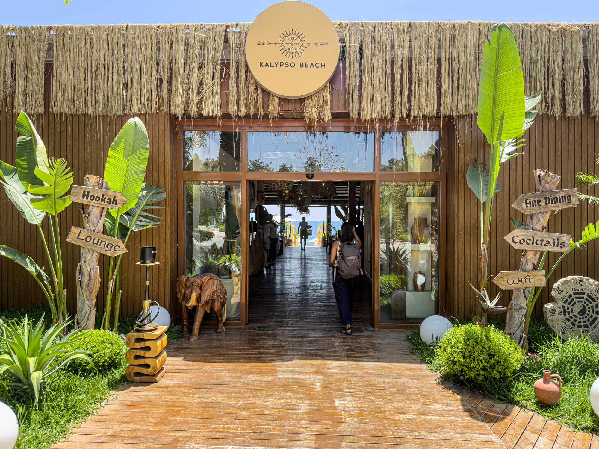 Entrance to Kalypso Beach club in Konyaaltı, with tropical decor, hanging grass canopy, and wooden signs pointing to lounge areas, cocktails, and wifi