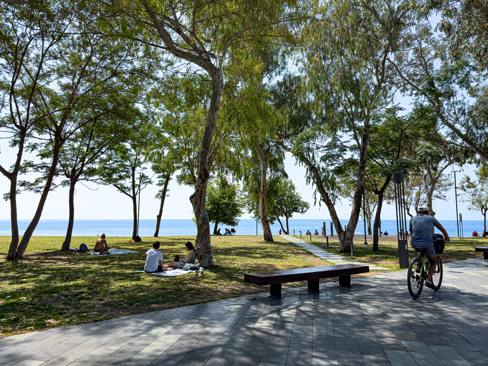 People relaxing and cycling under tree shade in Konyaaltı Beach Park, with the Mediterranean Sea in the background