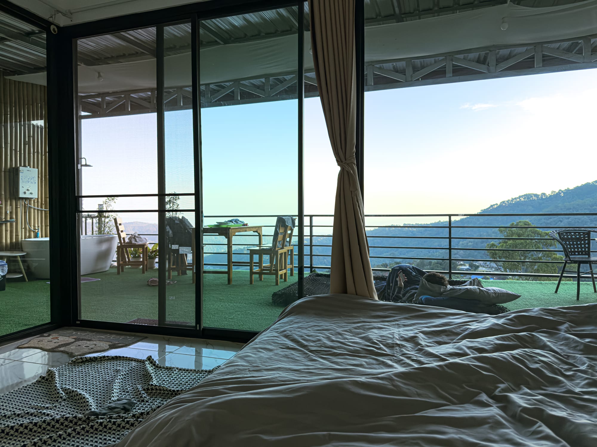View from inside a room at Mankhao Homestay showing a bed, balcony seating, and sweeping mountain scenery in Mae Rim