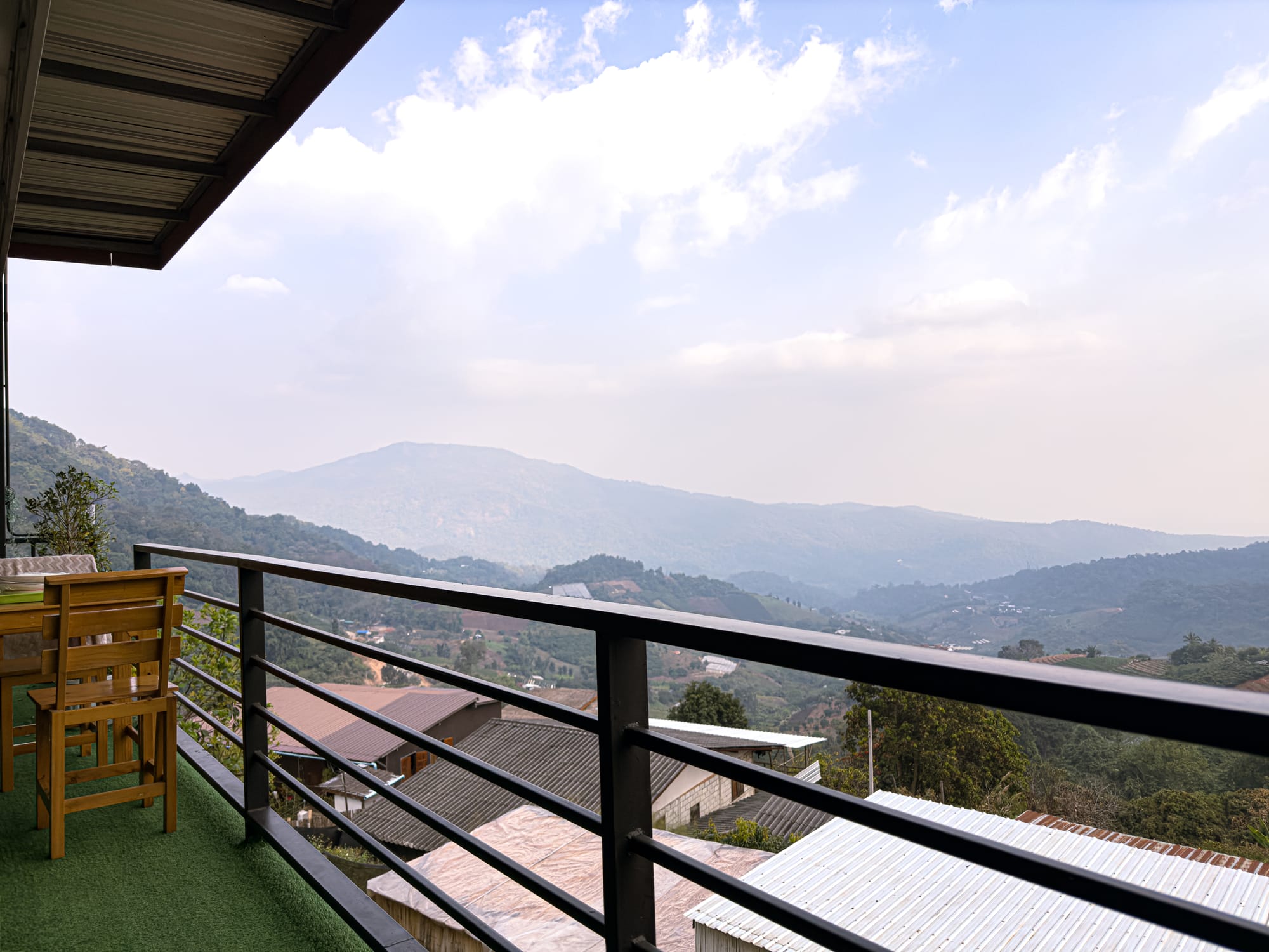 A wooden table and chairs on a balcony at Mankhao Homestay, with sweeping views of the surrounding green mountains and valleys in Mae Rim under a partly cloudy sky