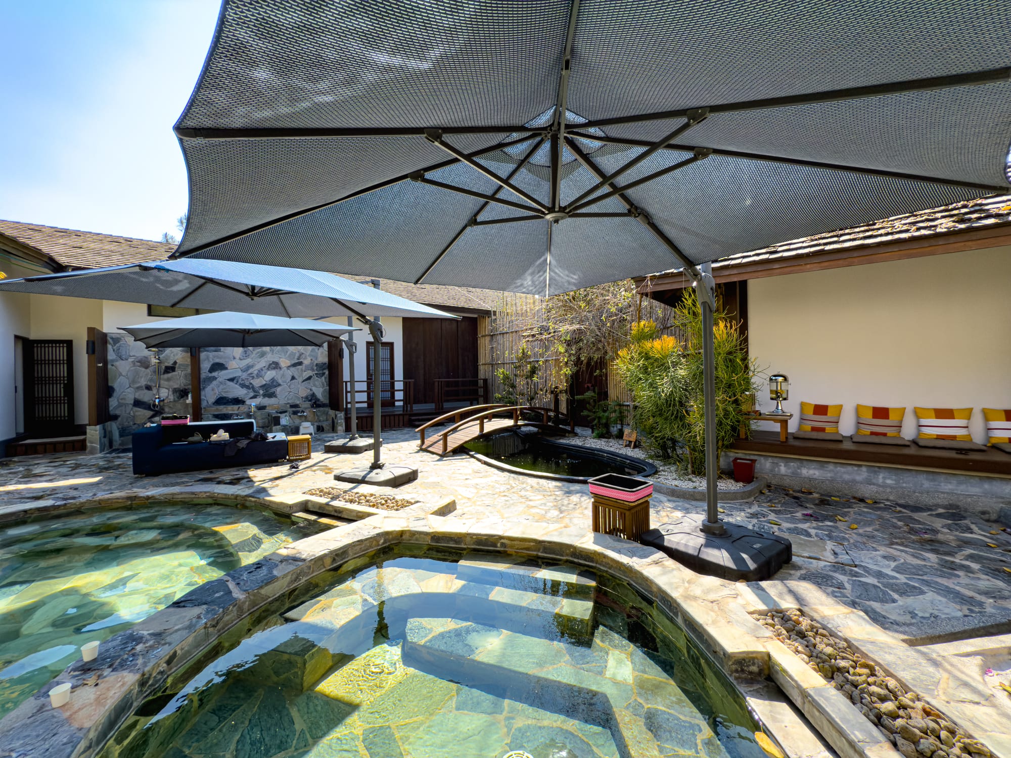 Stone onsen pools with umbrellas, seating, and a small wooden bridge at Onsen at Moncham