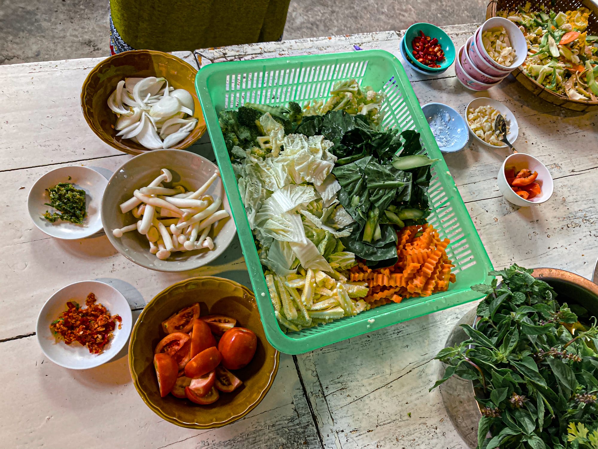 A colorful spread of fresh vegetables and herbs for May Kaidee’s vegan cooking class, including carrots, cabbage, leafy greens, mushrooms, tomatoes, chilies, and aromatic herbs arranged in bowls and baskets on a rustic white table