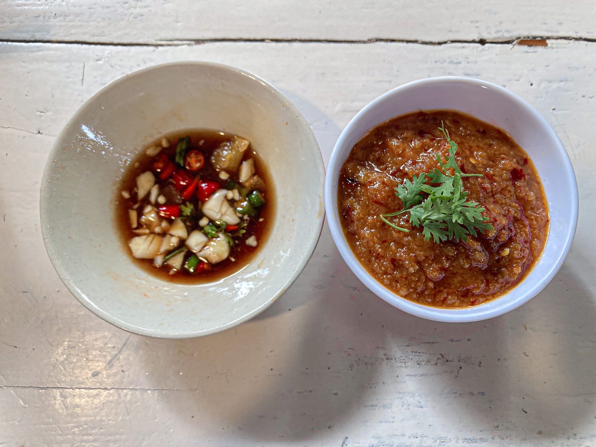 Overhead view of two small bowls on a rustic white table, one holding a soy-based dipping sauce with chopped garlic and red and green chilies, and the other containing a thick, reddish-brown roasted chili paste garnished with a sprig of cilantro