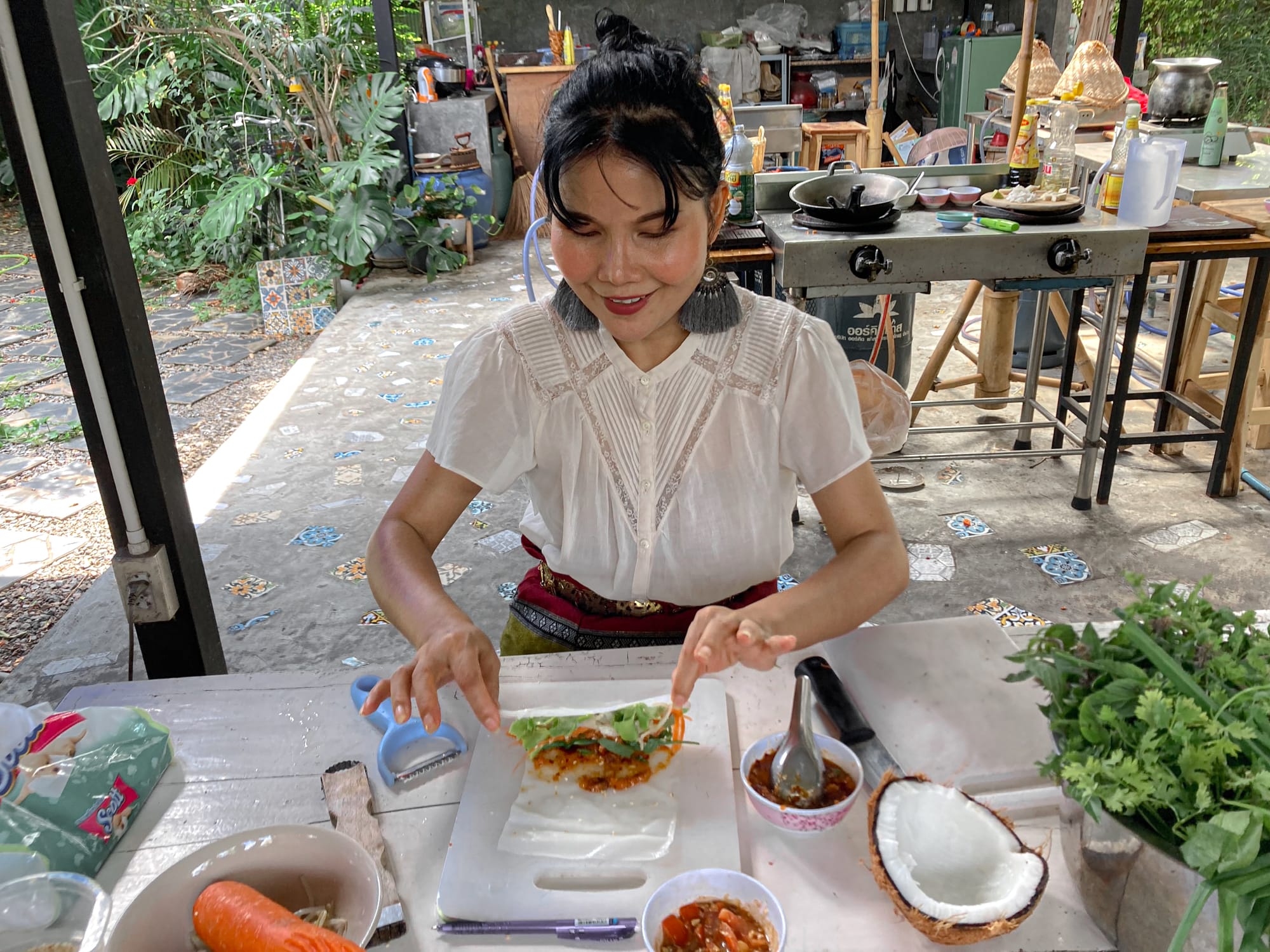 A cooking instructor at May Kaidee’s vegan cooking class in Chiang Mai smiles as she carefully folds fresh vegetables into rice paper, preparing spring rolls at a table filled with herbs, coconut, and colorful ingredients