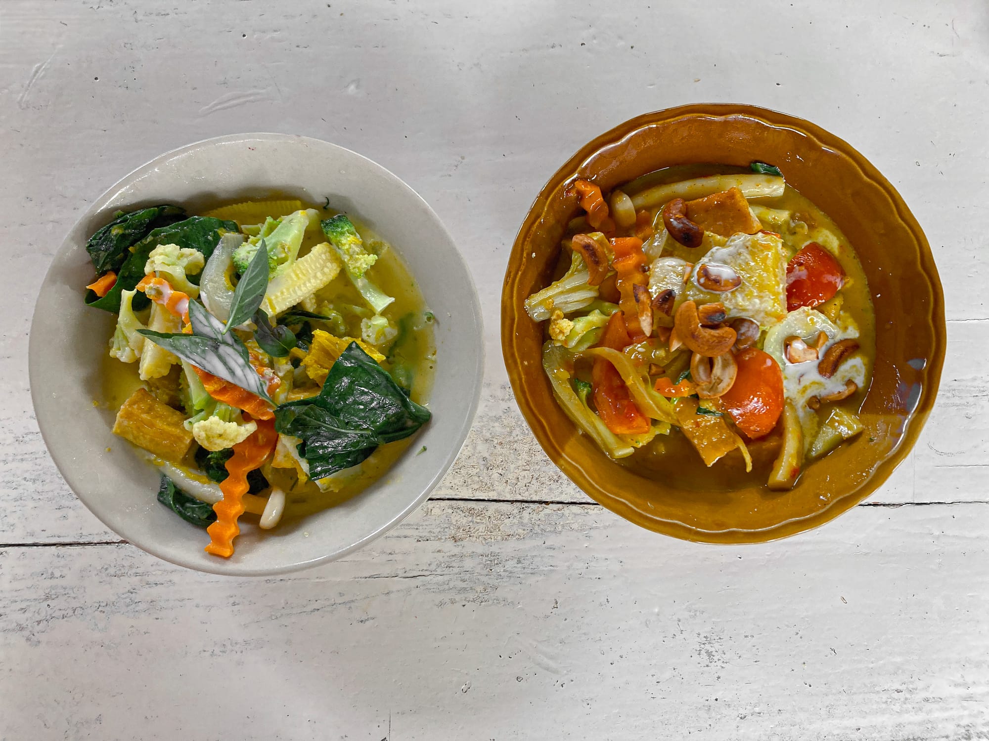 A white bowl of vegan green curry with broccoli, baby corn, carrots, and Thai basil sits beside a brown bowl of red curry filled with tofu, tomatoes, carrots, and cashews, both photographed on a rustic white wooden table