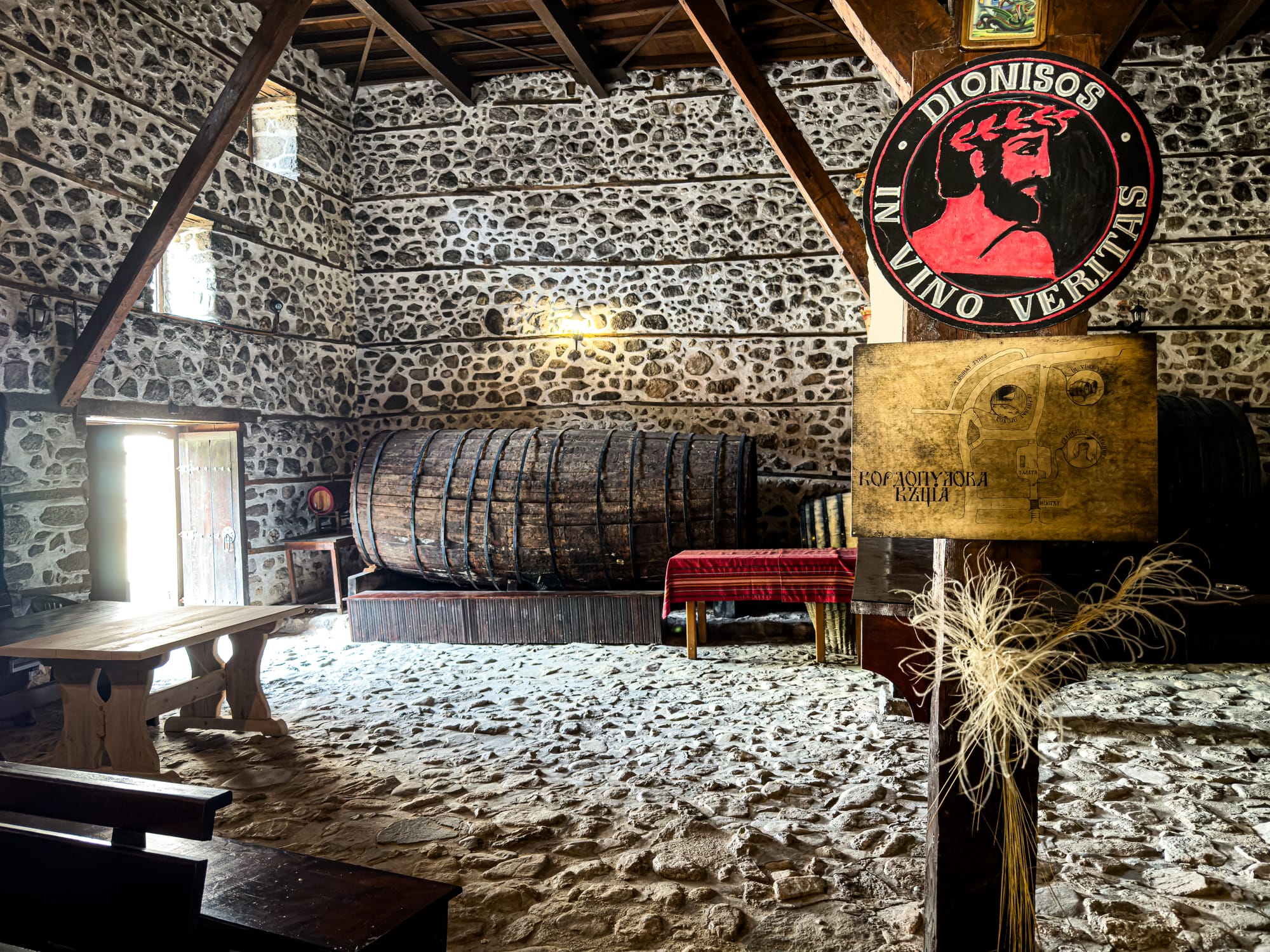 Interior of the Kordopulov House wine cellar in Melnik, Bulgaria, with stone walls, wooden beams, a massive old wine barrel, and a painted sign of Dionysos reading ‘In Vino Veritas'