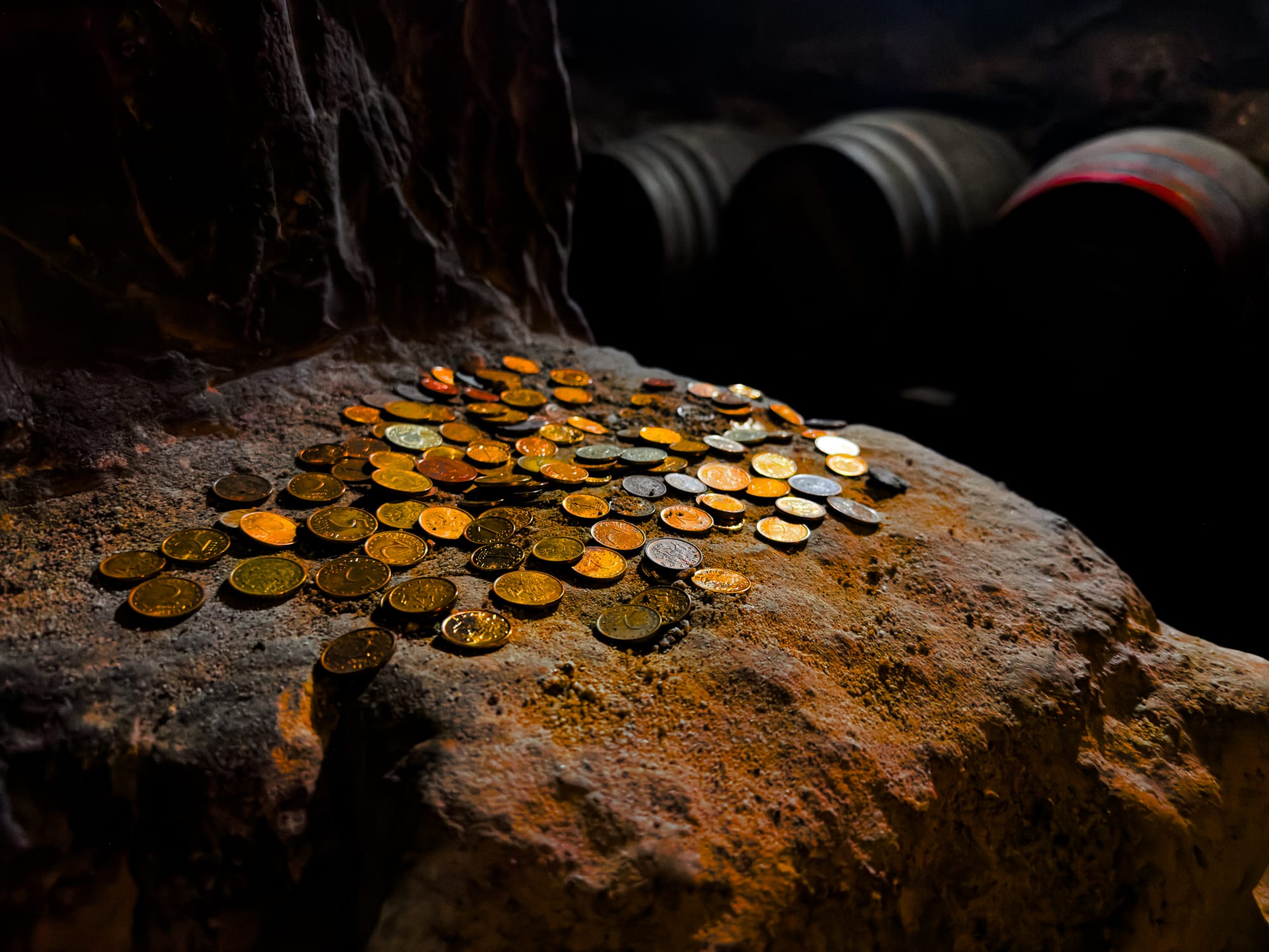 ile of coins scattered on a rough stone surface inside a dimly lit Melnik wine cellar, with wine barrels visible in the background