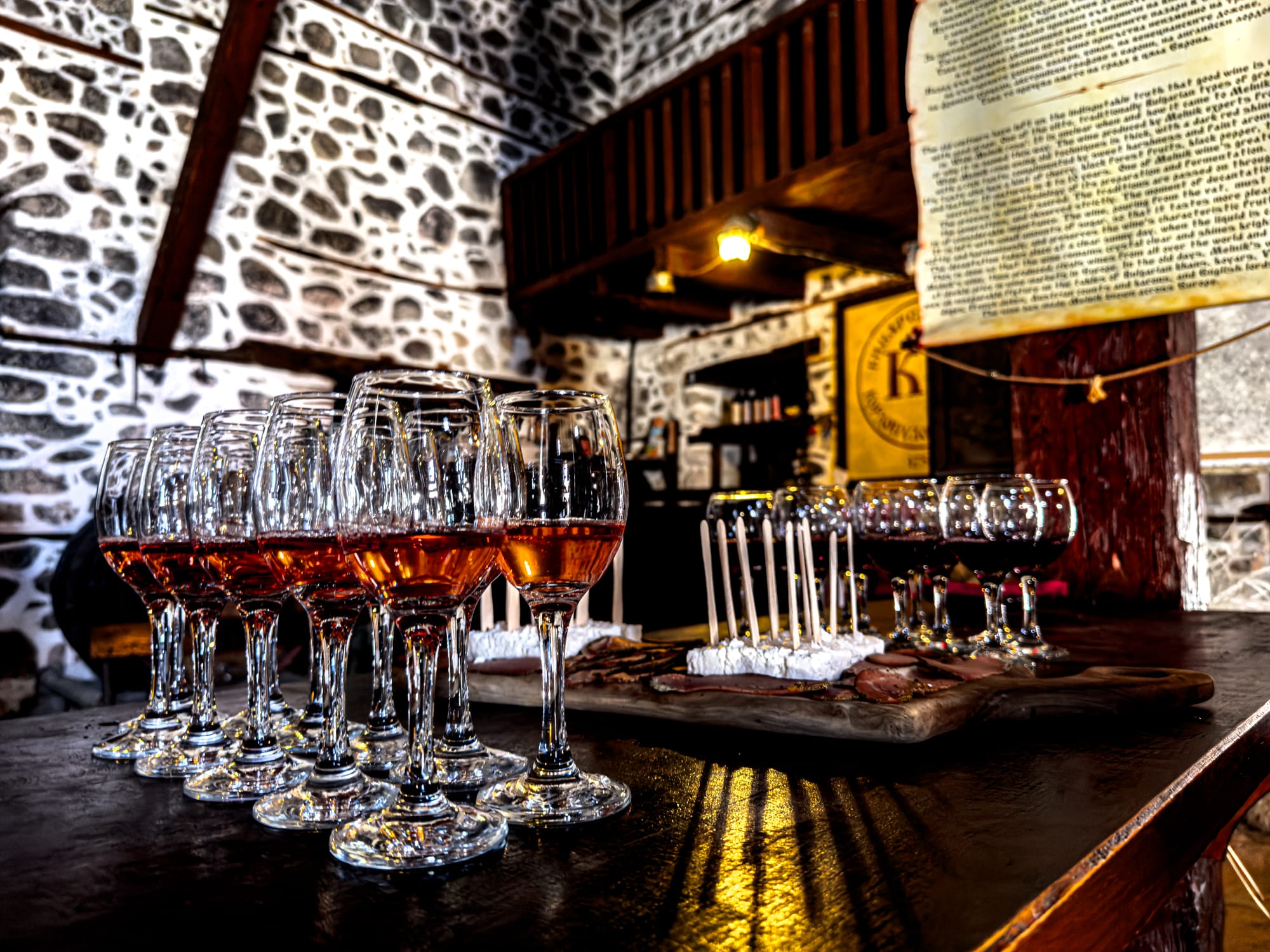 Close-up of glasses of rosé wine arranged on a wooden table in a stone-walled winery in Melnik, Bulgaria, with a platter of cheeses and cured meats in the background