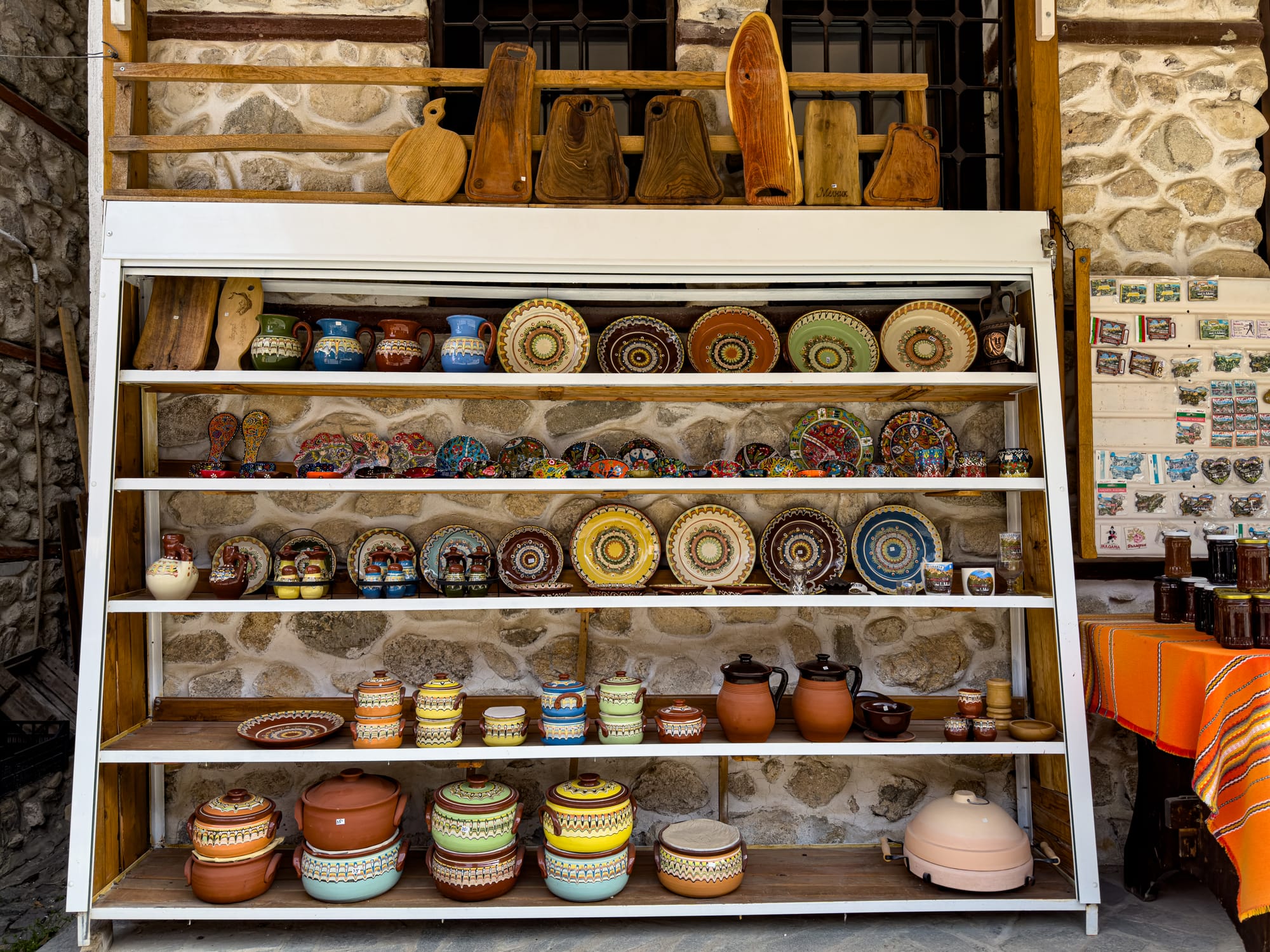 Display shelves in Melnik, Bulgaria, filled with colorful traditional pottery, plates, and clay cooking pots, with cutting boards and souvenirs arranged around a stone wall