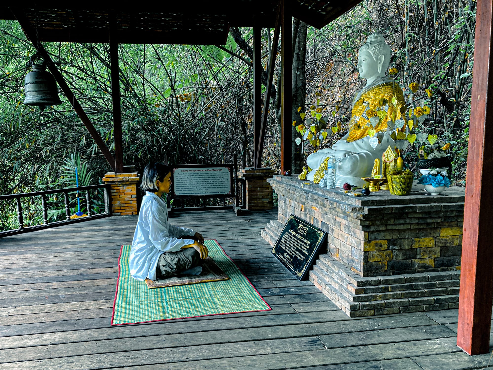A woman in white clothing kneels on a woven mat, meditating in front of a white Buddha statue draped in a golden sash, surrounded by offerings and golden leaf ornaments, at Wat Pha Lat along the Monk’s Trail in Chiang Mai