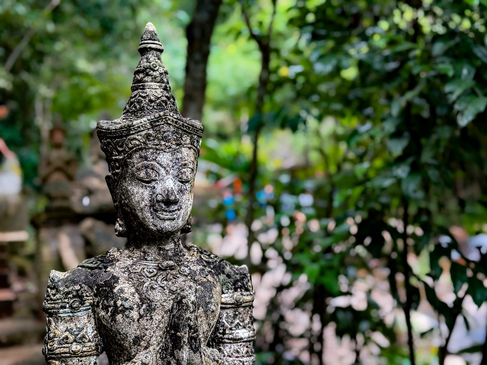 Close-up of an aged stone guardian statue with intricate carvings and a peaceful face, set against a blurred backdrop of dense jungle foliage at Wat Pha Lat on the Monk’s Trail