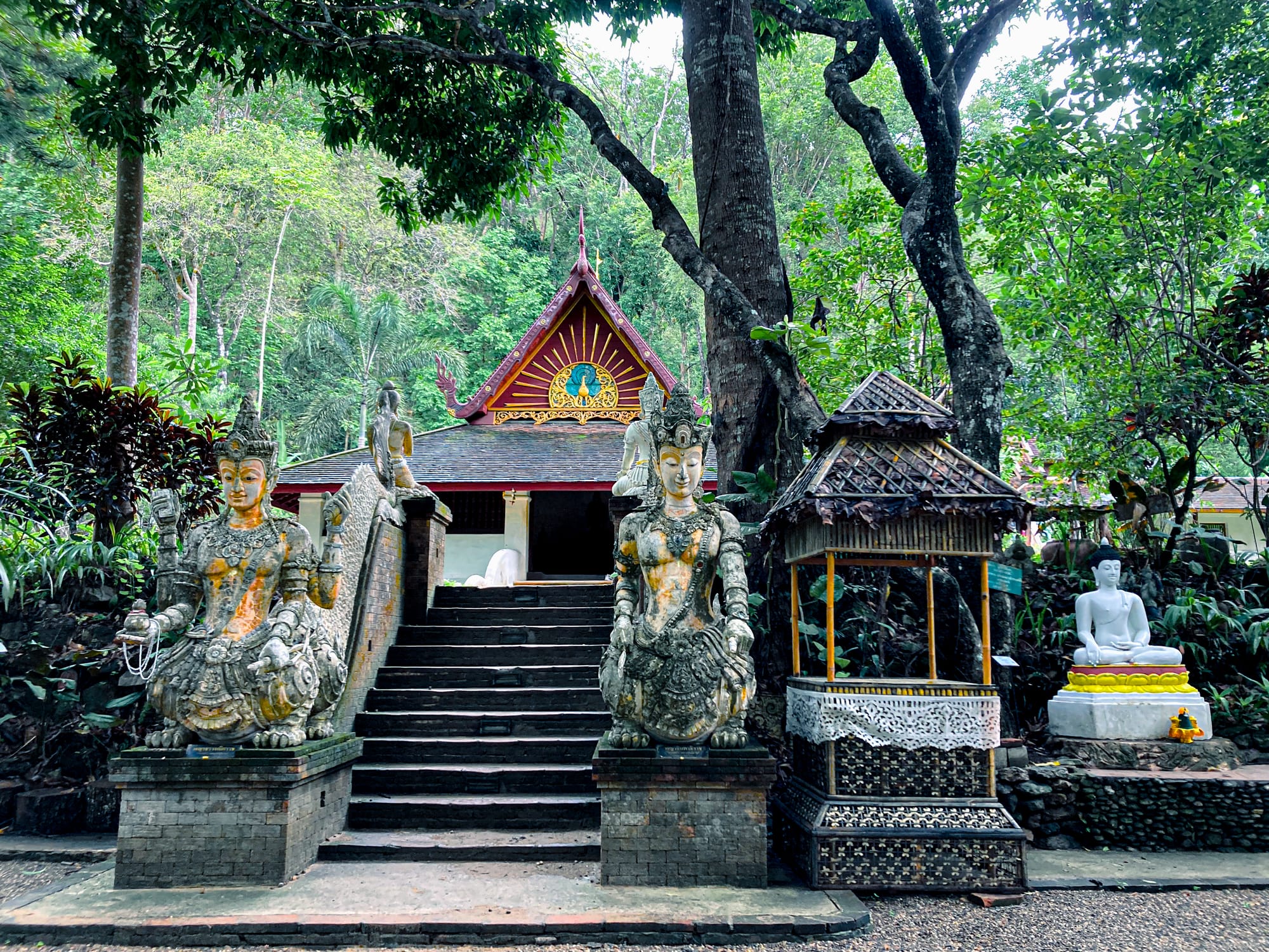 Two intricately carved guardian statues flank the staircase to a forest temple at Wat Pha Lat, with a gabled roof decorated in gold and red, and a serene Buddha statue seated nearby under tall jungle trees