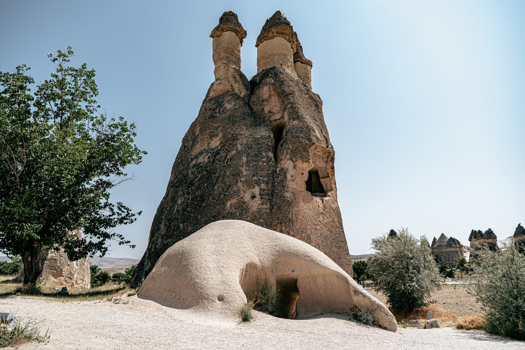 A large, twin-capped fairy chimney in Paşabağ (Monks Valley), Cappadocia, featuring carved windows and a smaller rock formation with a low entrance at its base, surrounded by trees and dry landscape under a clear blue sky