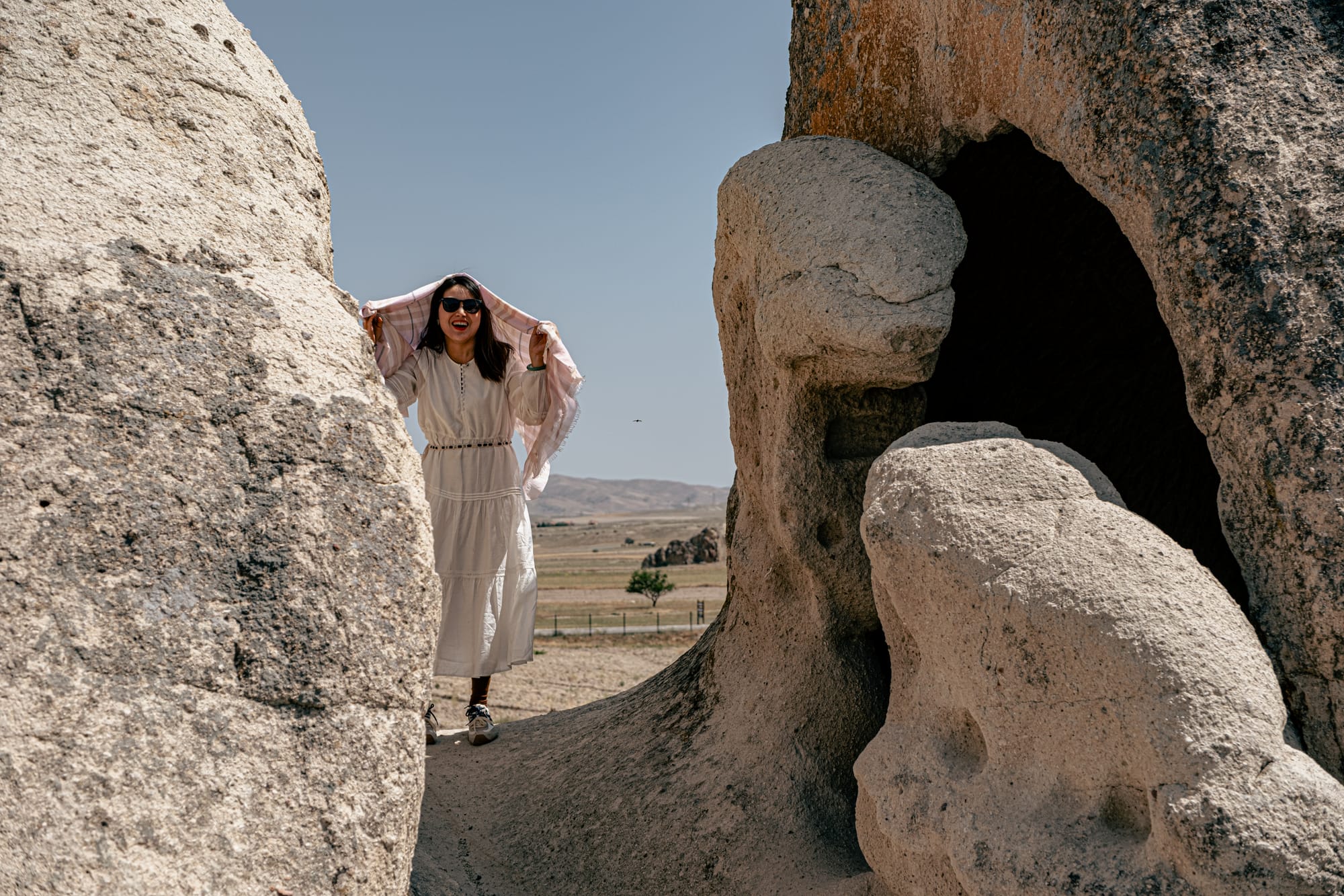A woman in a white dress and sunglasses, holding a scarf above her head, walks between large volcanic rock formations in Paşabağ (Monks Valley), Cappadocia, with open plains and hills in the background