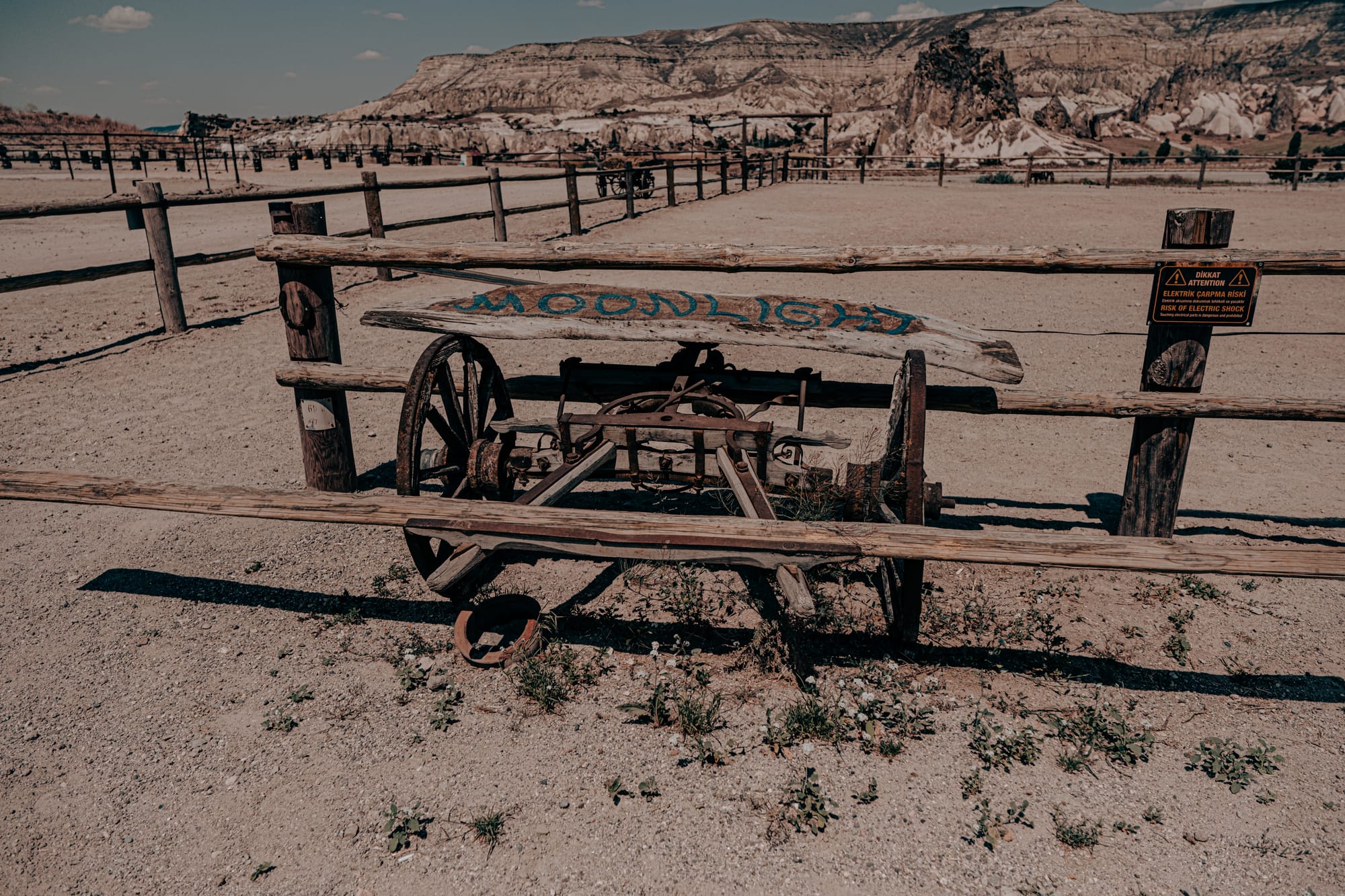 Rustic wooden cart with “Moonlight” painted on its plank, set within a fenced area at Moonlight Ranch in Cappadocia, with panoramic views of the region’s rocky mountains and open arena