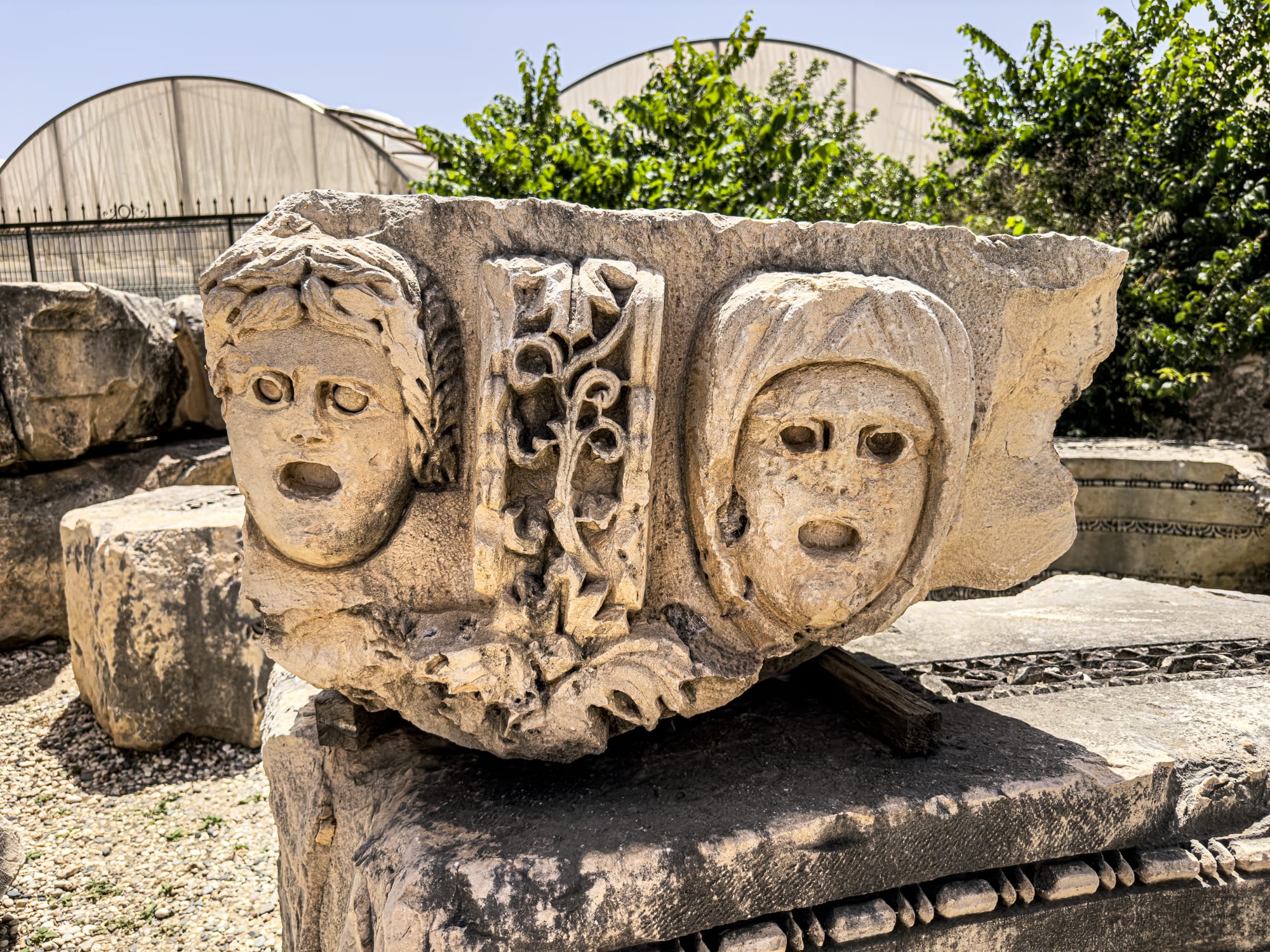 Ancient Roman-era stone carving of theatrical masks at Myra archaeological site near Demre, Turkey, with greenhouse domes and vegetation in the background