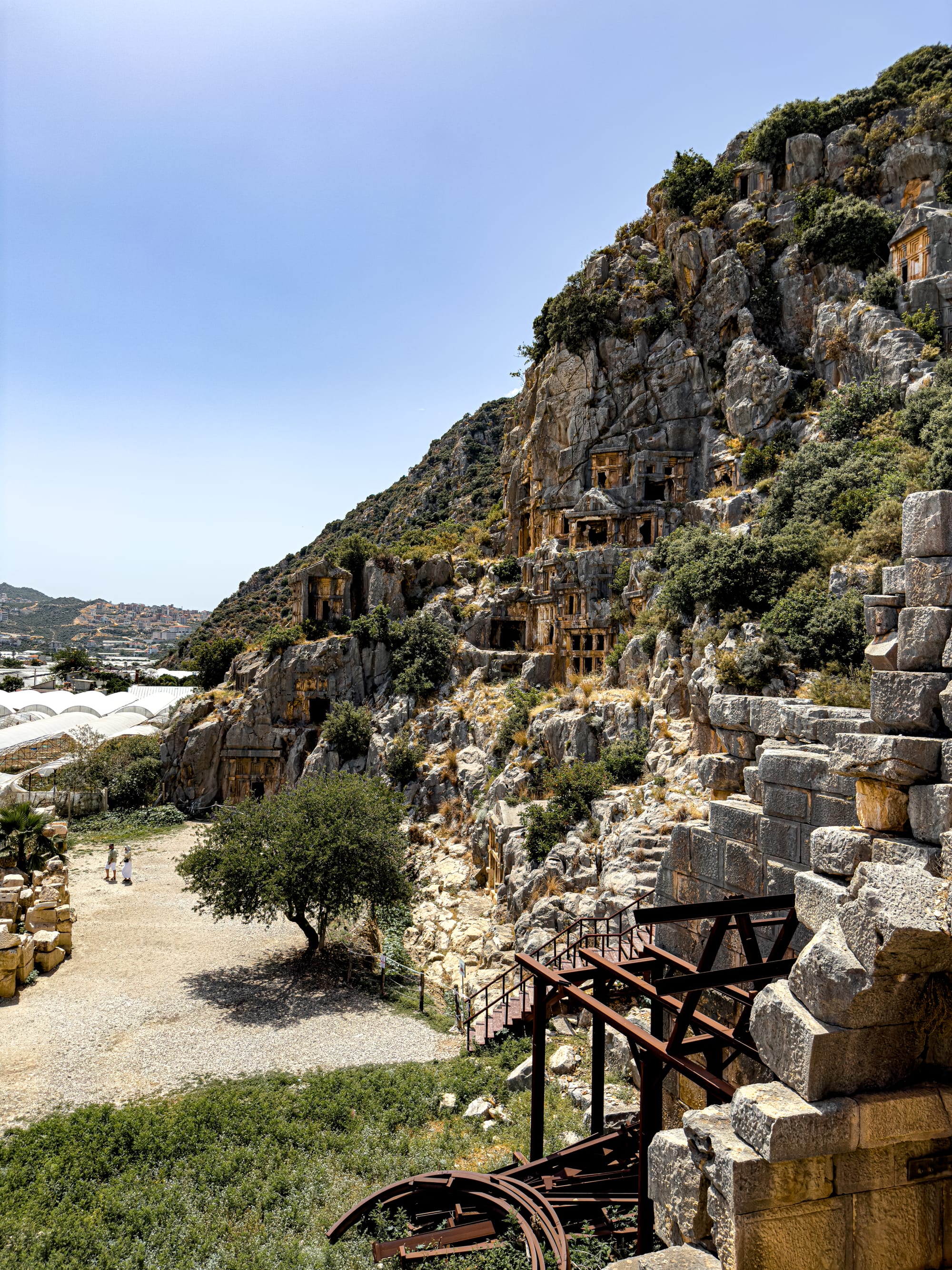 Lycian rock-cut tombs at the ancient city of Myra in Demre, seen from the Roman theater with foreground ruins and a tree-lined visitor path below