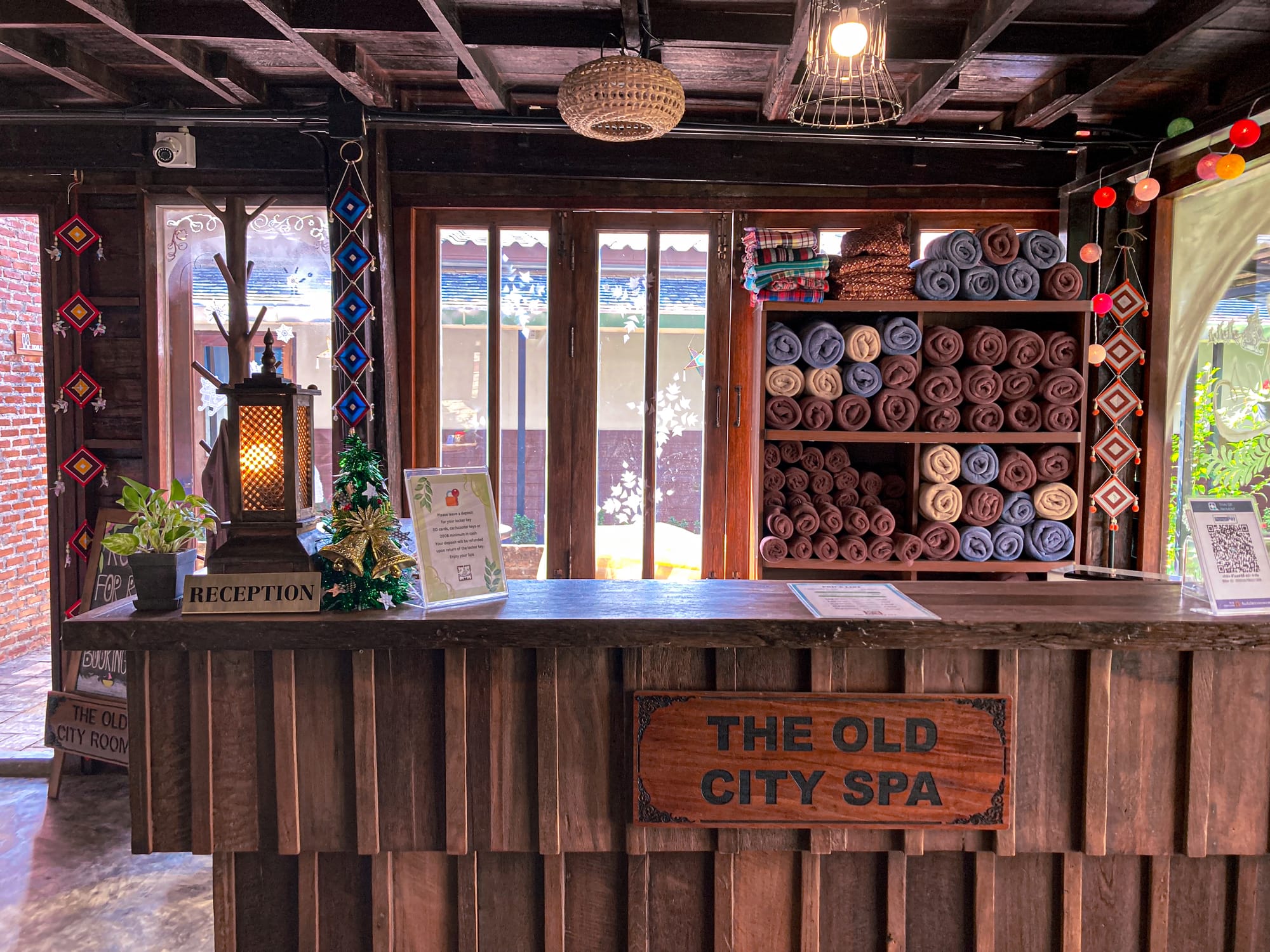 Reception area at Old City Spa in Chiang Mai with a wooden desk, lantern, colorful hanging decorations, and shelves filled with rolled towels
